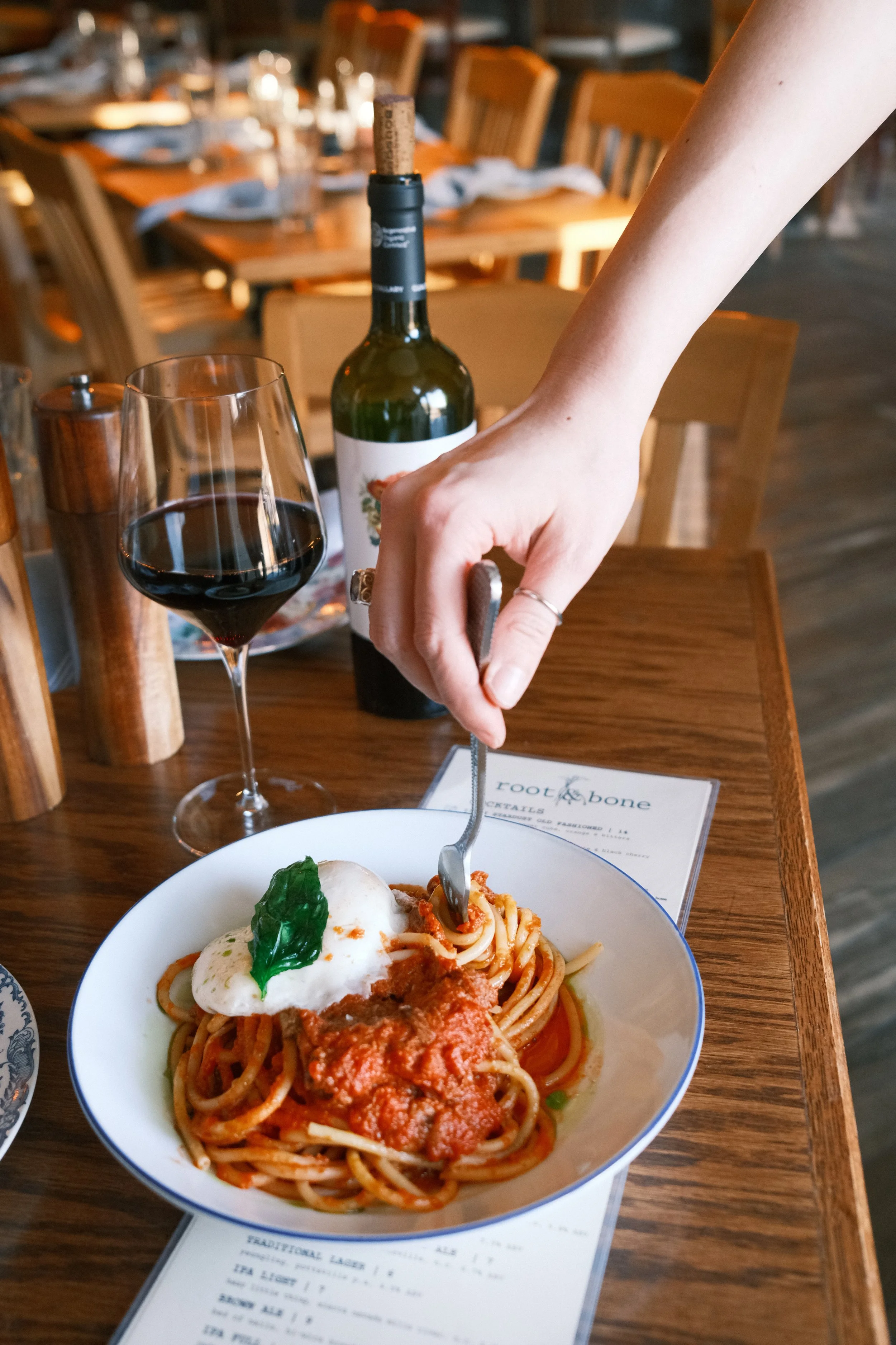 A person is cutting into a plate of spaghetti with tomato sauce, topped with a poached egg and basil leaf. A glass of red wine and a bottle of wine are on the table, which also has a wooden pepper grinder and a wine menu.