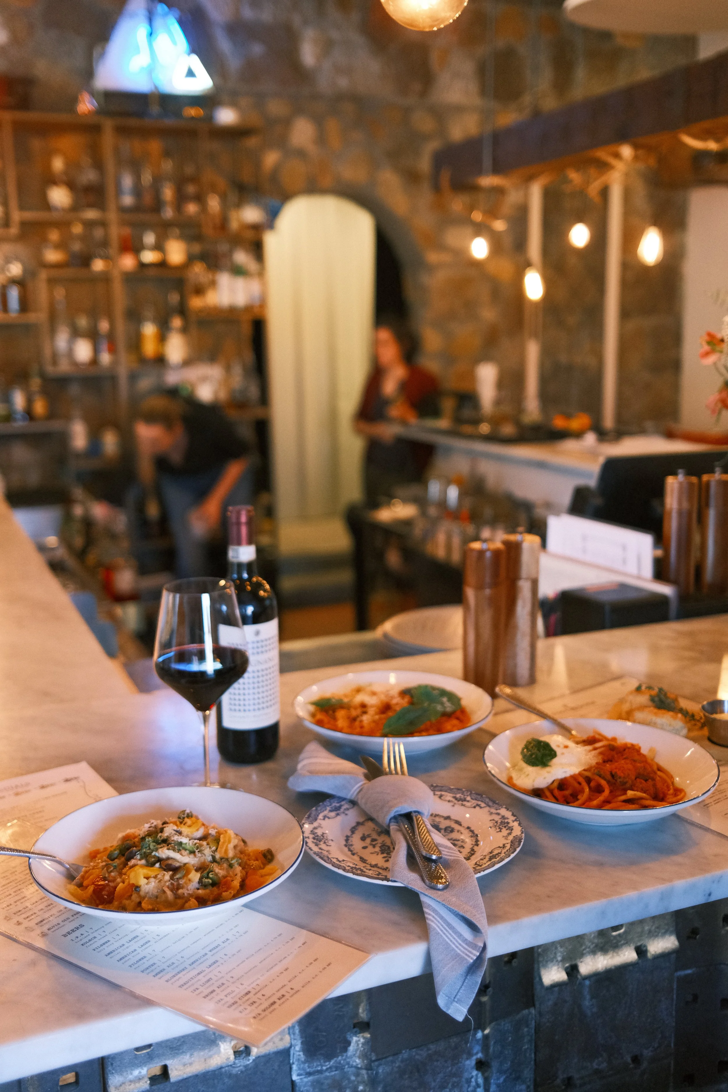 A marble countertop with three bowls of pasta, a glass of red wine, a bottle of wine, and a napkin with silverware, in a cozy restaurant with warm lighting and brick walls.