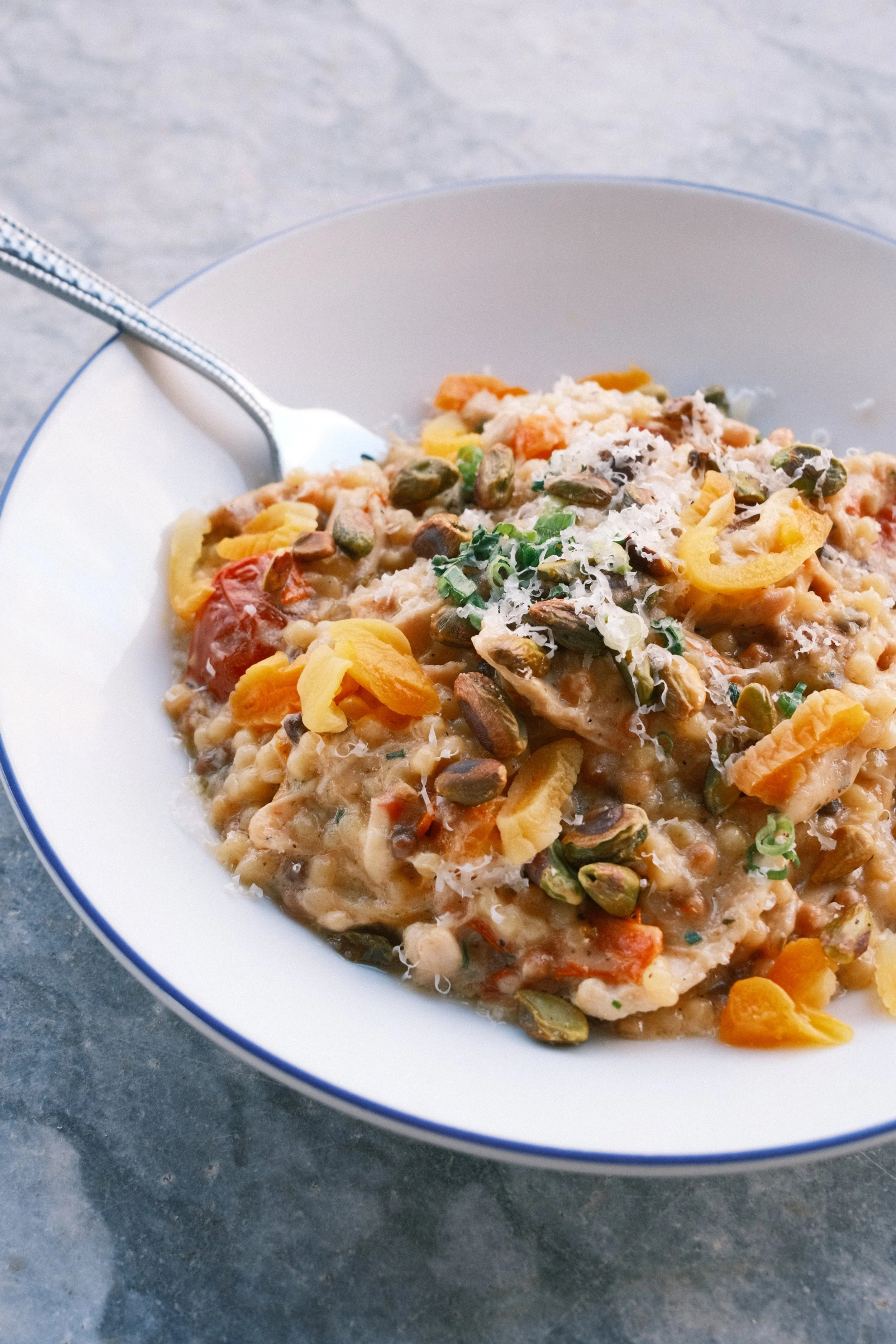 A bowl of hearty vegetable and grain stew topped with shredded cheese and chopped green onions, served with a fork on a gray surface.