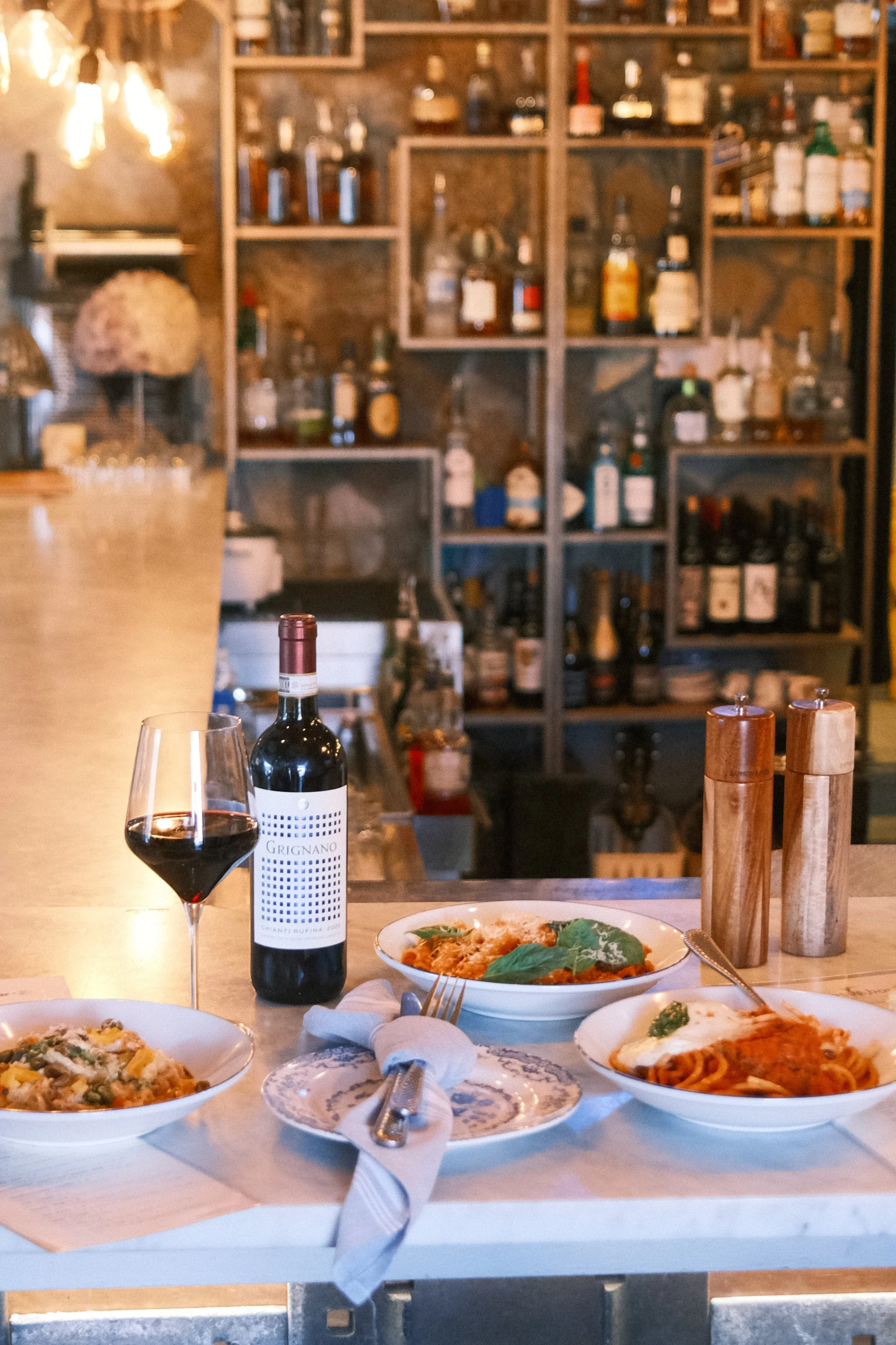 A dining table with a bottle of red wine, a glass of red wine, three plates of pasta, and salt and pepper shakers in a cozy restaurant with a bookshelf filled with bottles and jars in the background.