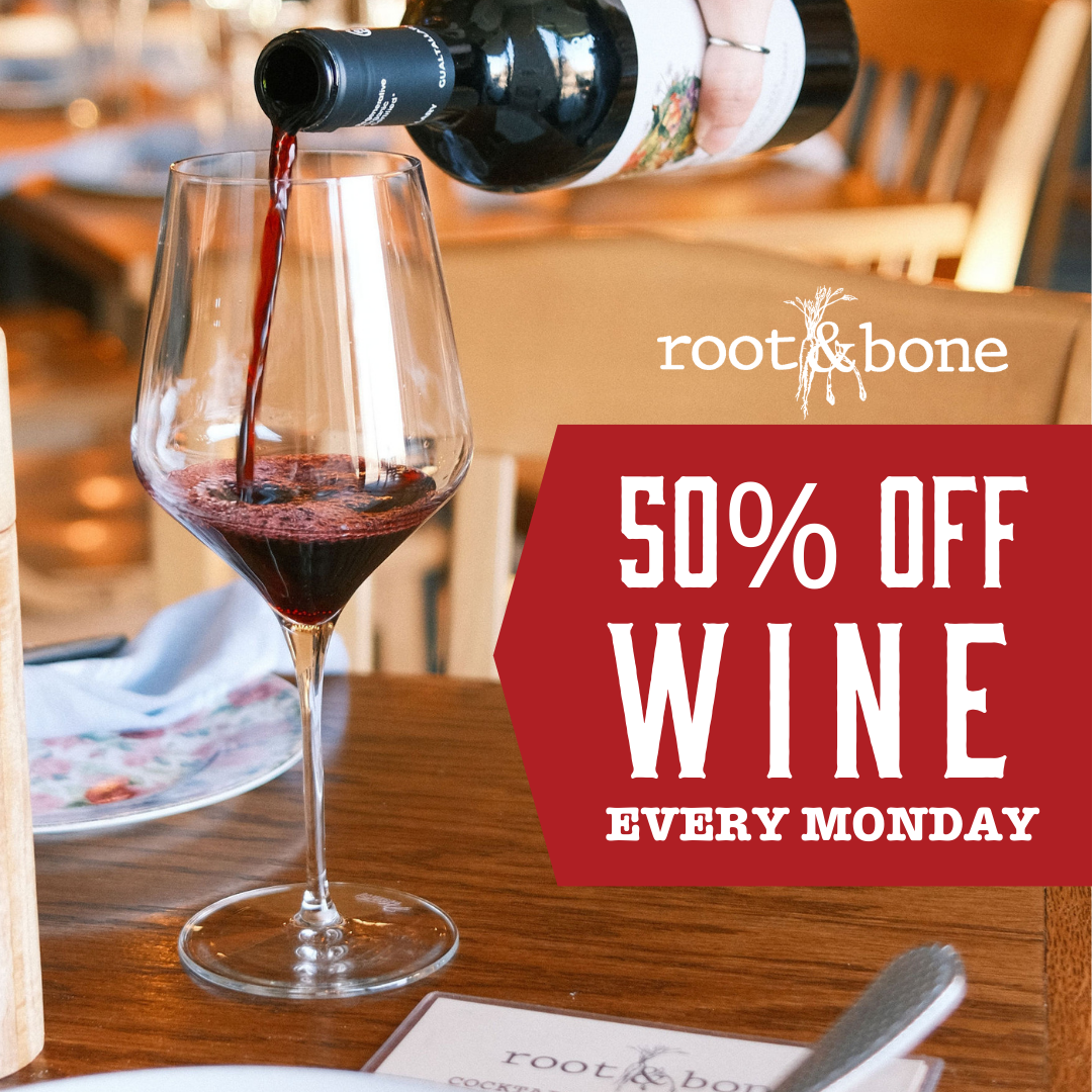A glass of red wine being poured from a bottle on a wooden table in a restaurant setting, with a promotional sign offering 50% off wine every Monday.