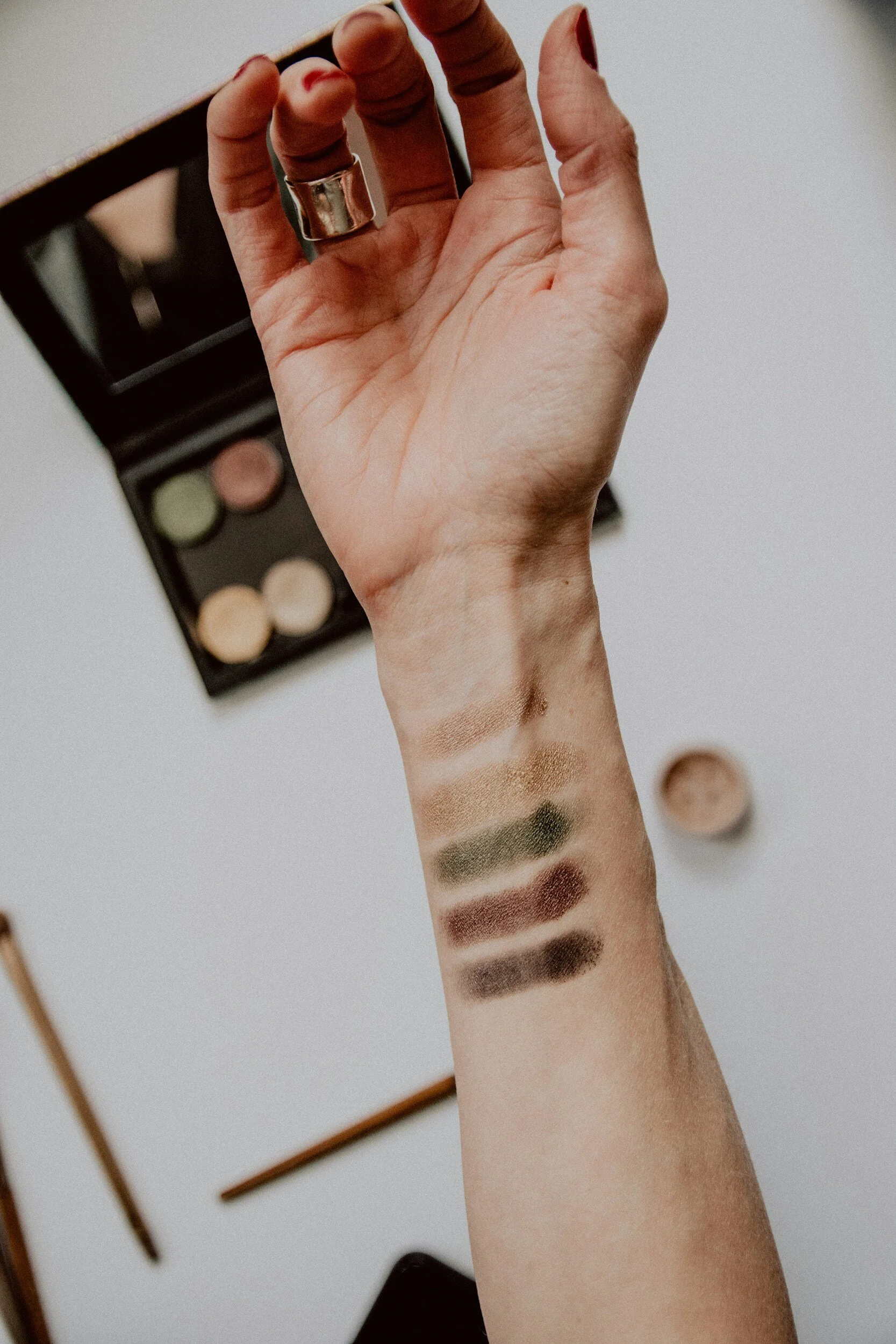 A person's arm with swatches of eyeshadow in various shades, with an eyeshadow palette in the background.