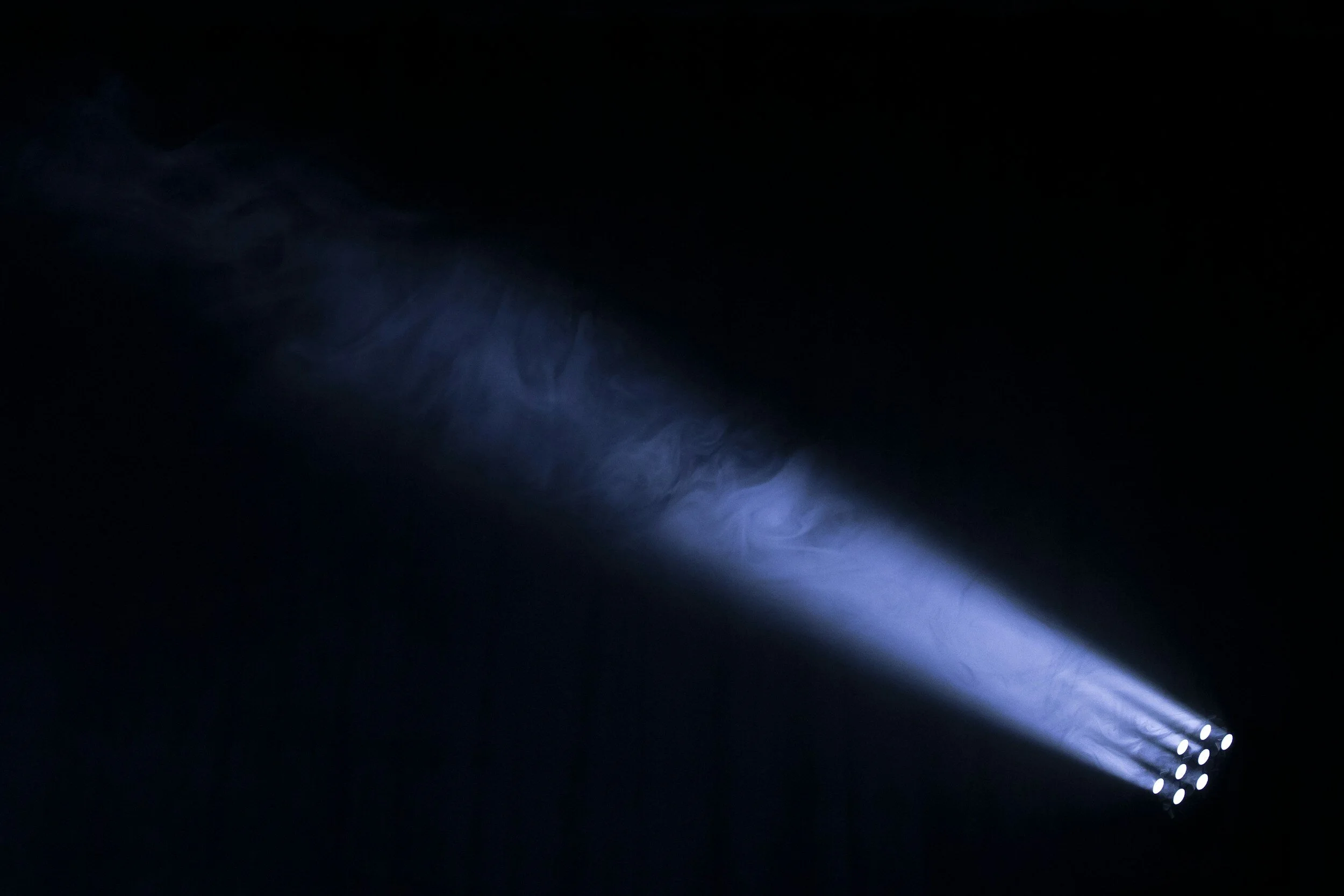 A dark scene with a beam of blueish white light shining diagonally from a multi-LED light source, creating a gradient of illumination in a smoky or foggy atmosphere.
