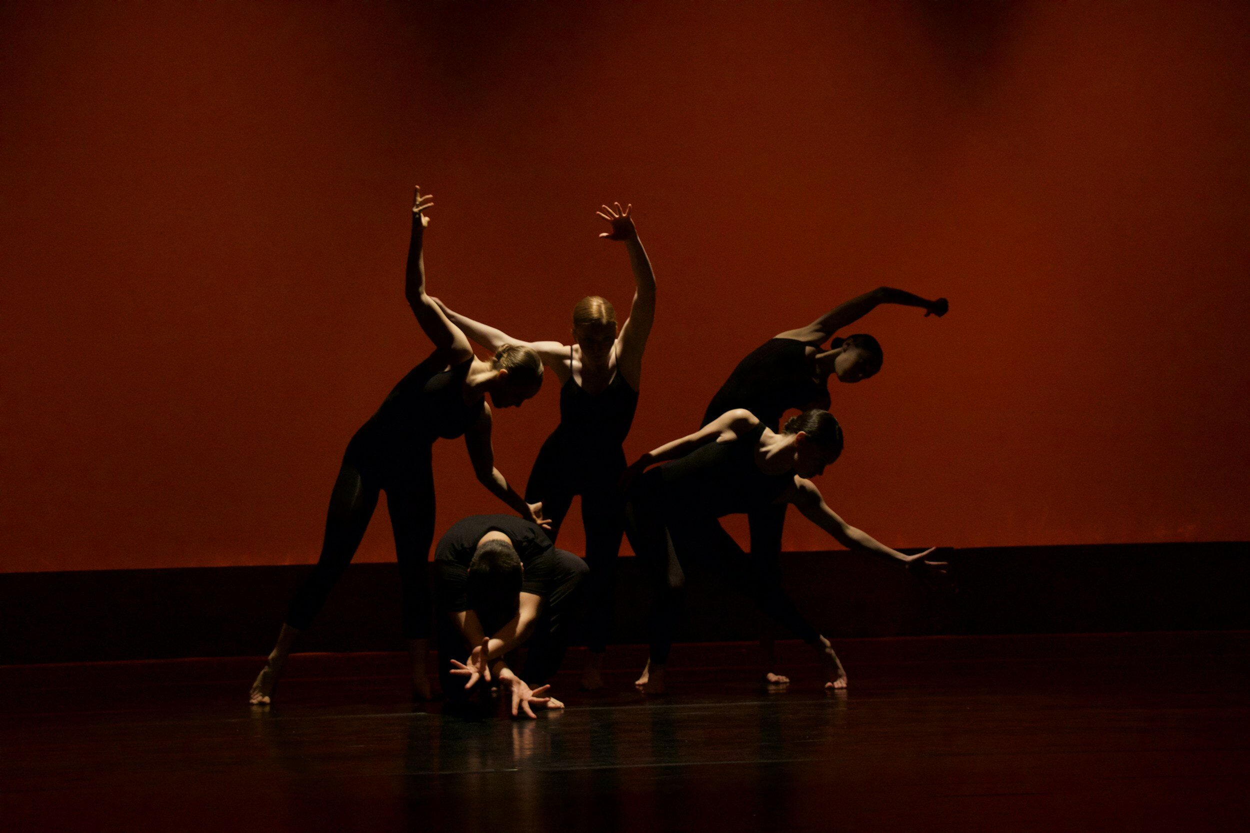 Five dancers performing a contemporary dance on stage with a dark background. They are dressed in black and positioned in various dynamic poses.