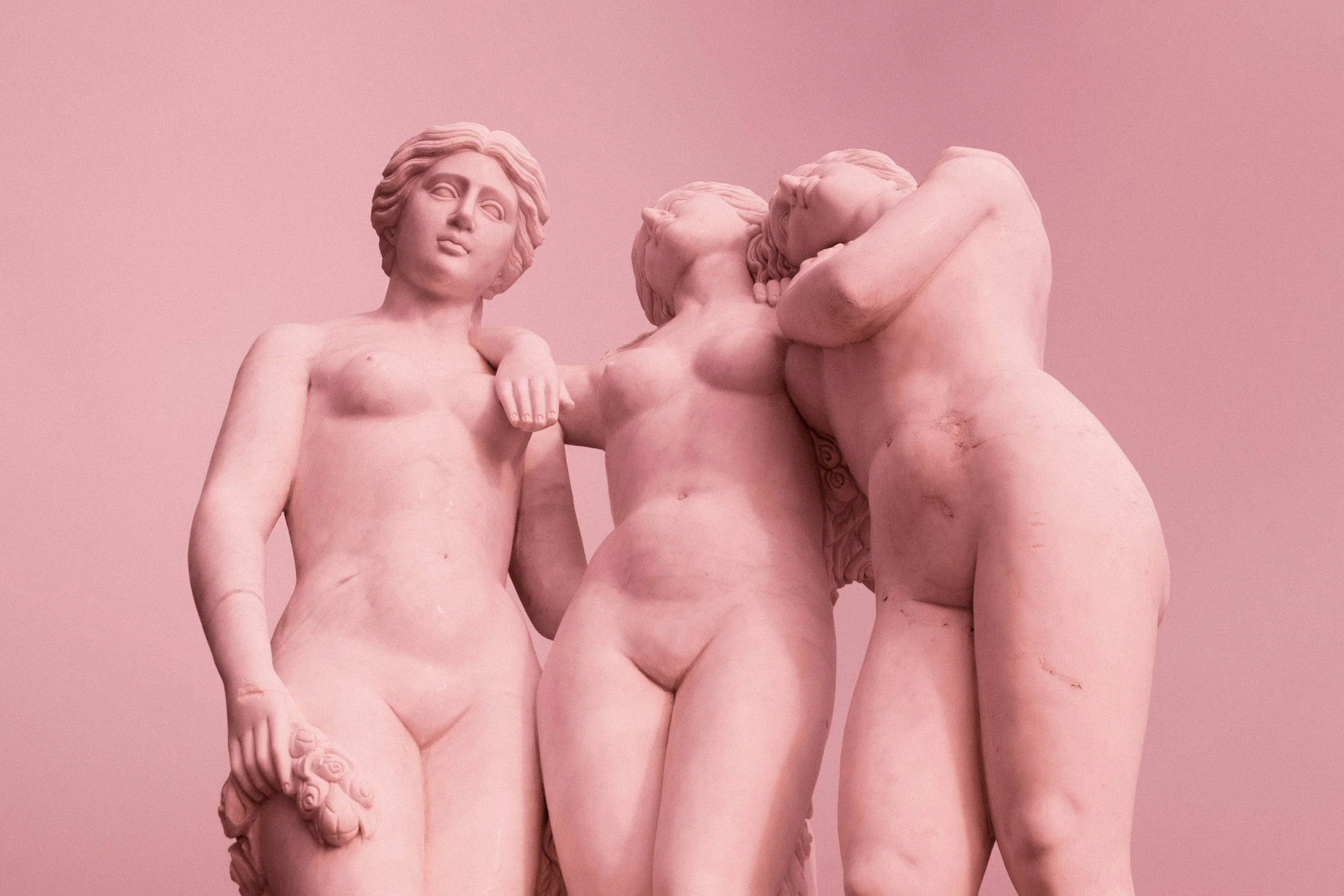 Marble sculpture of three nude figures with intertwined arms and facial expressions, set against a pink background.