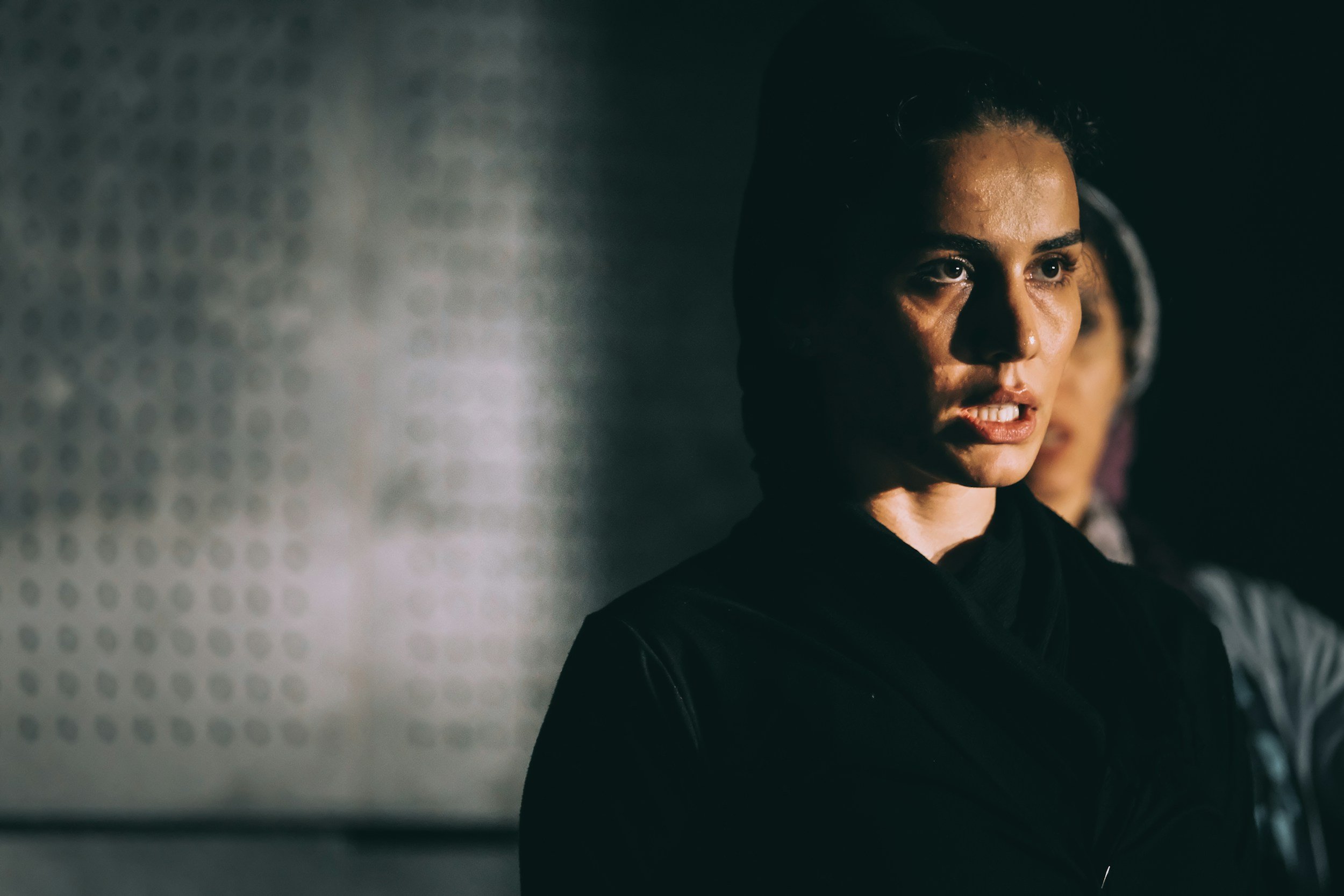 A woman with dark hair and a serious expression standing in a dimly lit setting, with a blurred background.