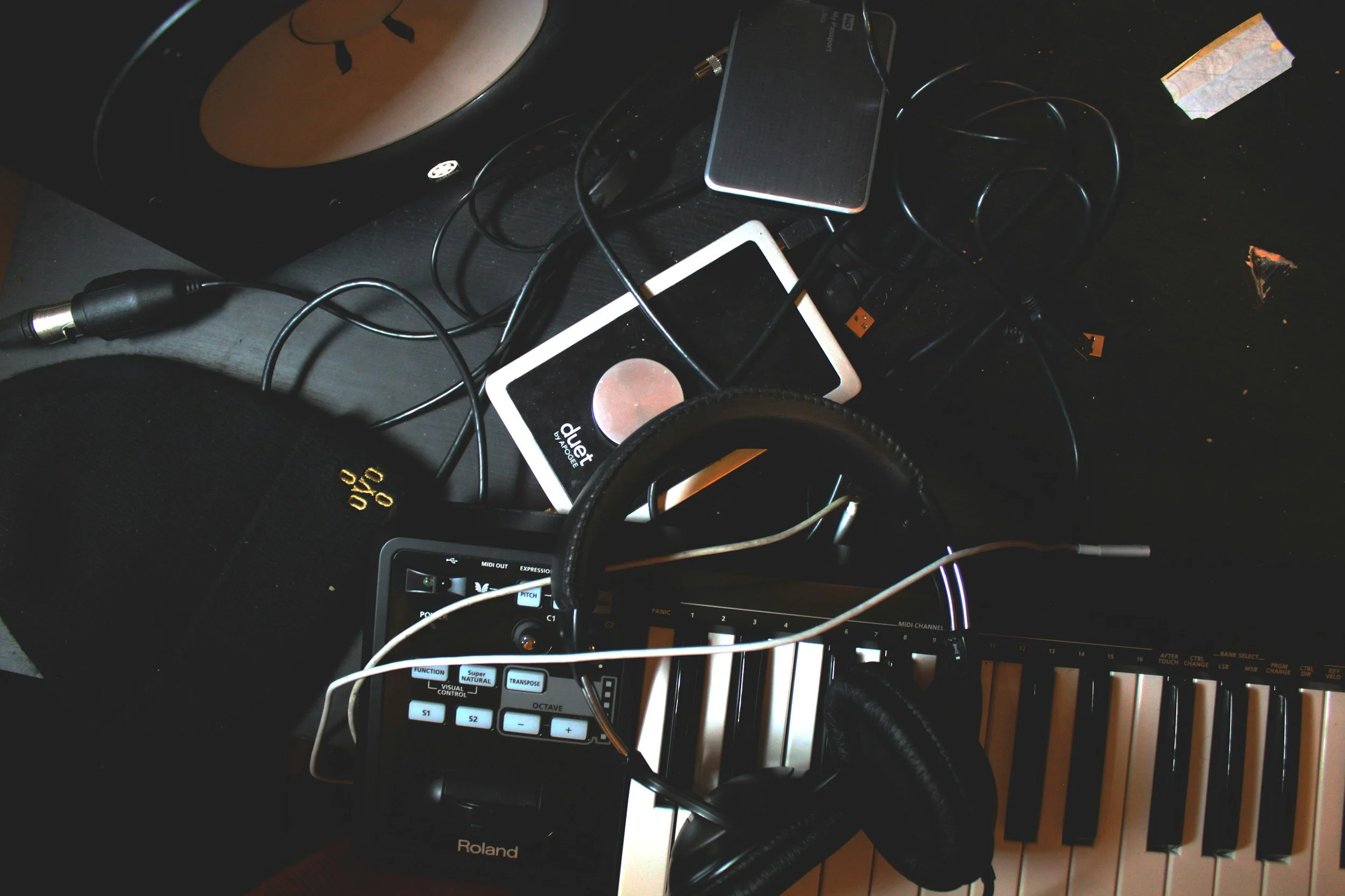 A cluttered music production workspace with a keyboard, headphones, a drum pad, a smartphone, a speaker, and various cables on a black surface.