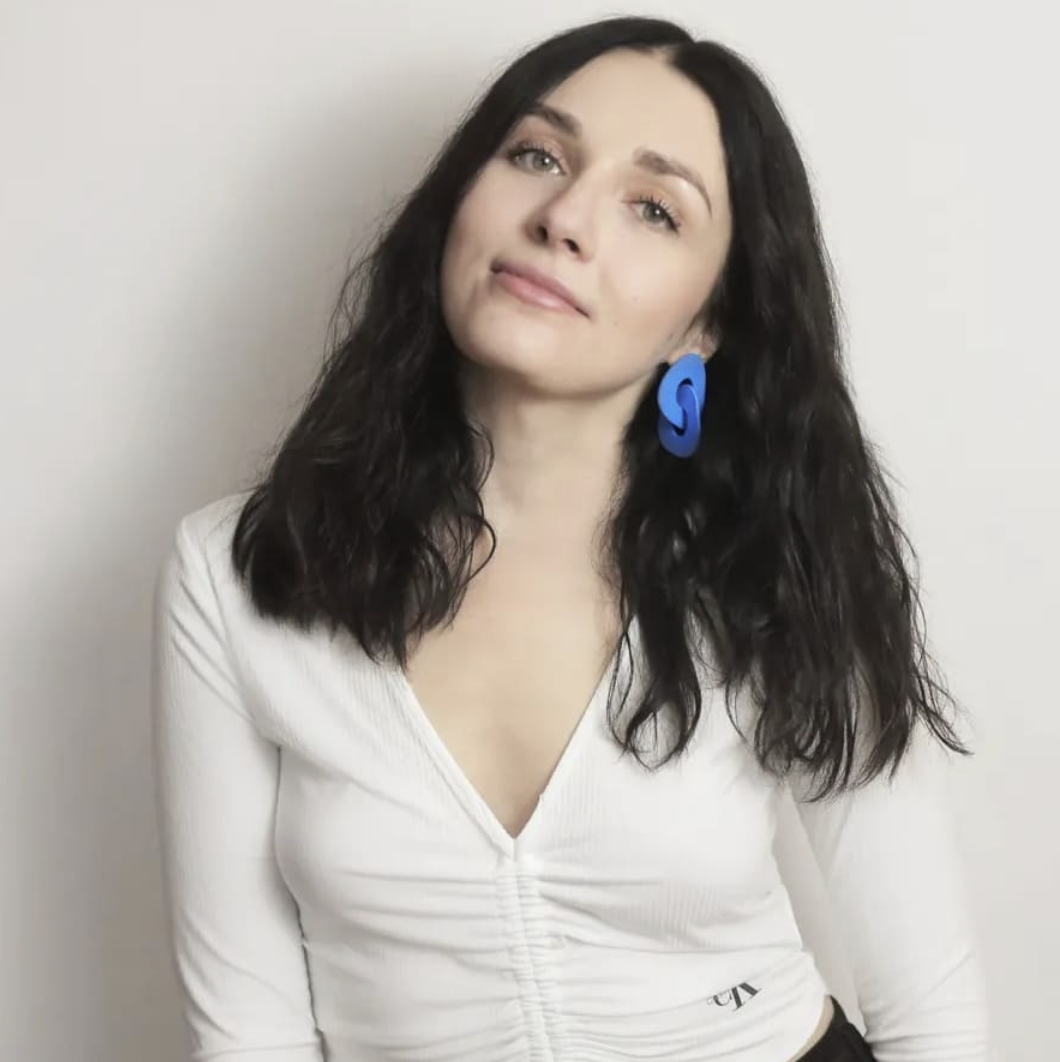 A woman with long dark curly hair, light skin, and blue earrings, wearing a white top, standing against a plain wall.