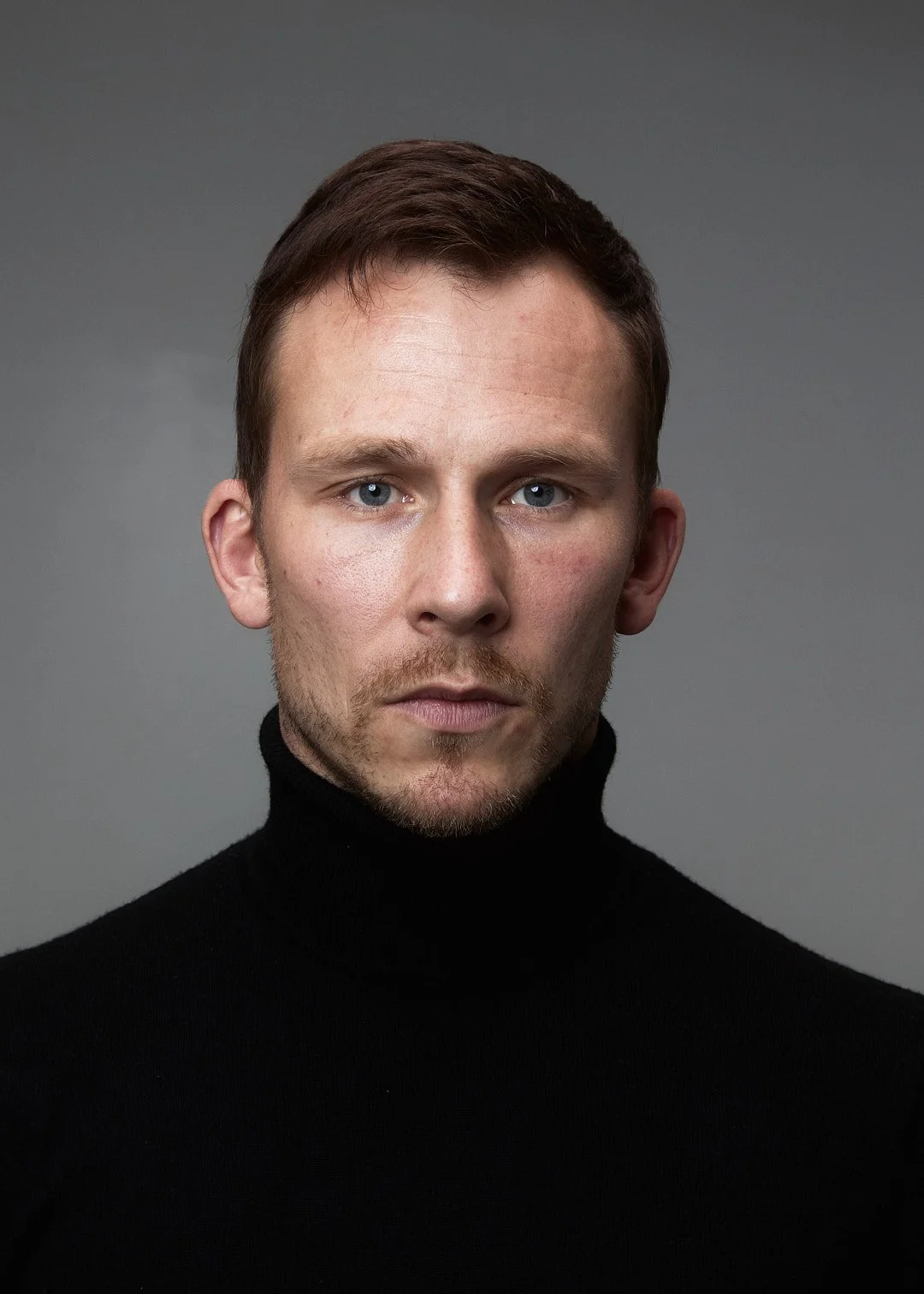 Portrait of a man with short brown hair, blue eyes, light skin, and light facial hair, wearing a black turtleneck sweater against a gray background.