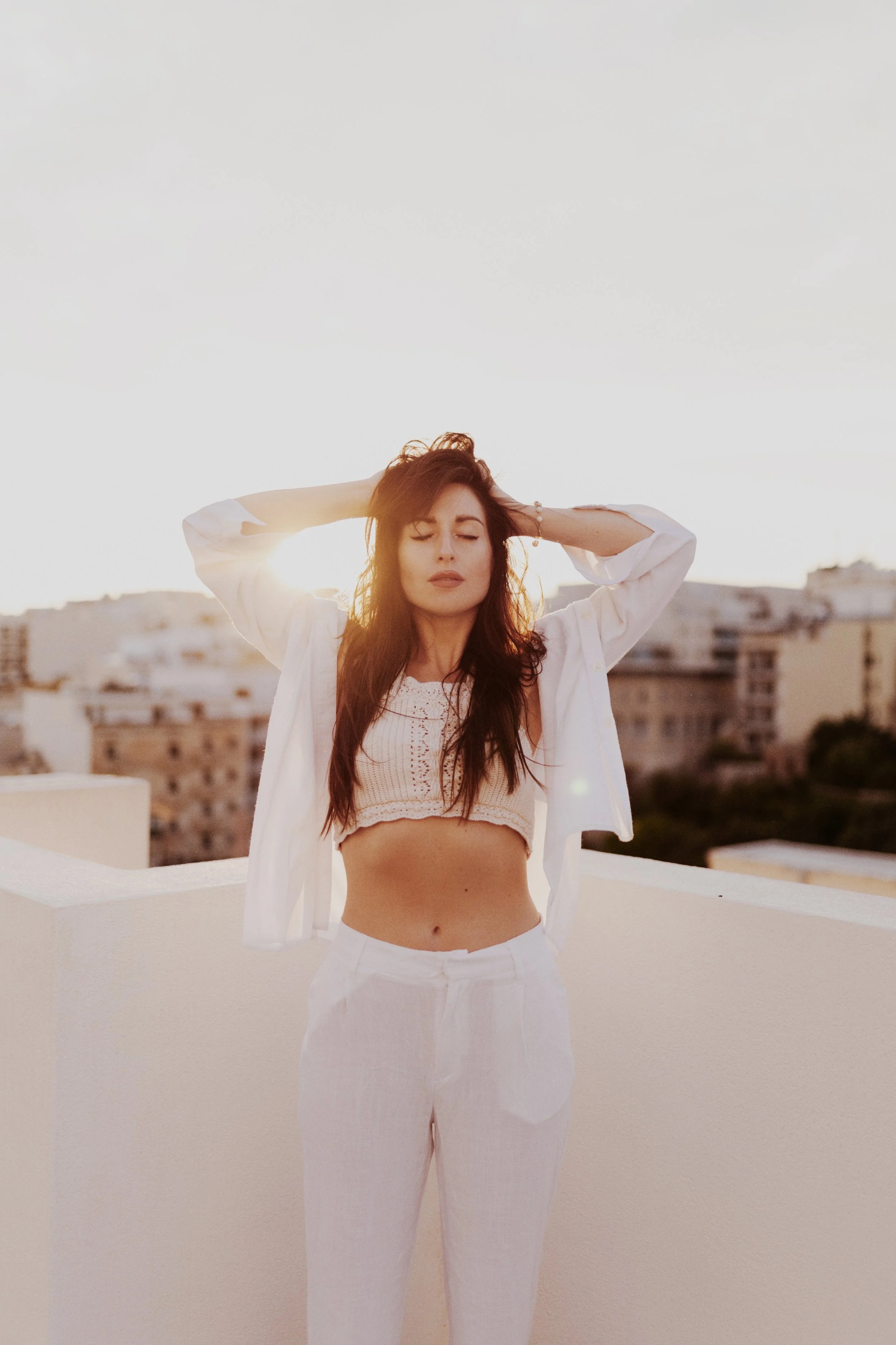 A woman with red hair, wearing a white cropped top and pants, standing on a rooftop at sunset with her eyes closed and hands in her hair.