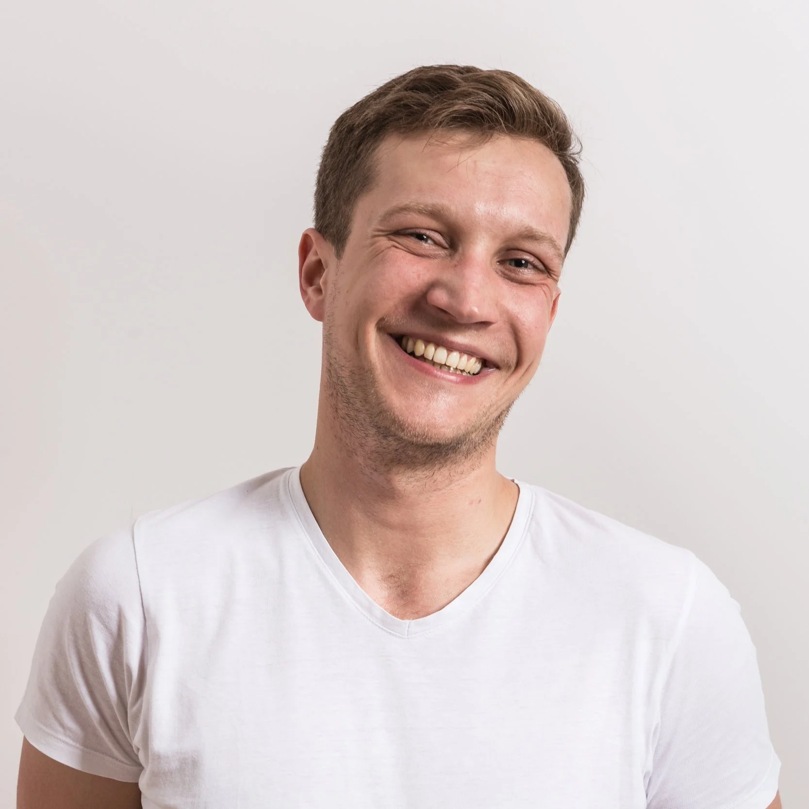 Young man with short brown hair smiling, wearing a white t-shirt, against a plain light gray background.