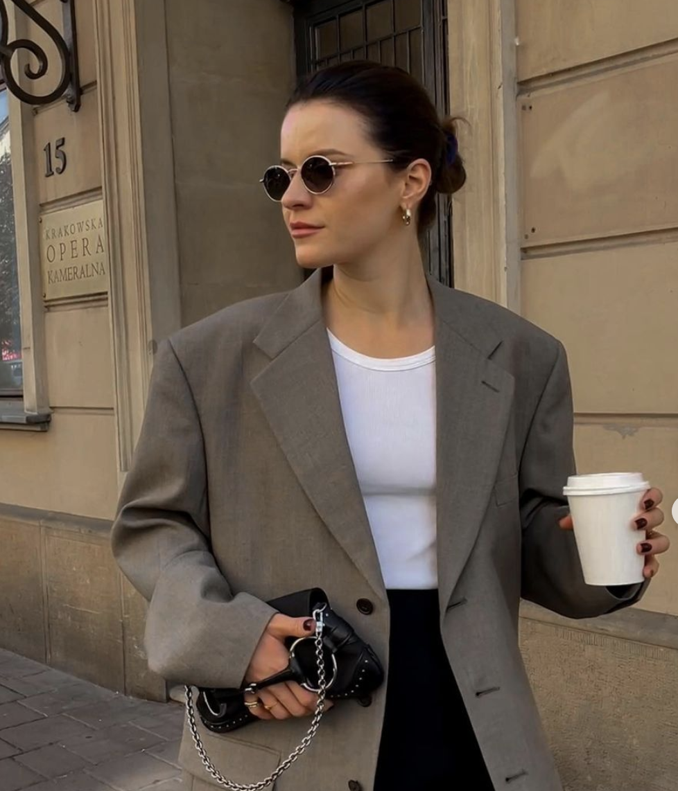 A woman in sunglasses, a gray blazer, and a white shirt holding a coffee cup and a black purse with chain strap, standing outside a building with a beige wall and a sign that reads 'Krakowska Opera Kameralna'.