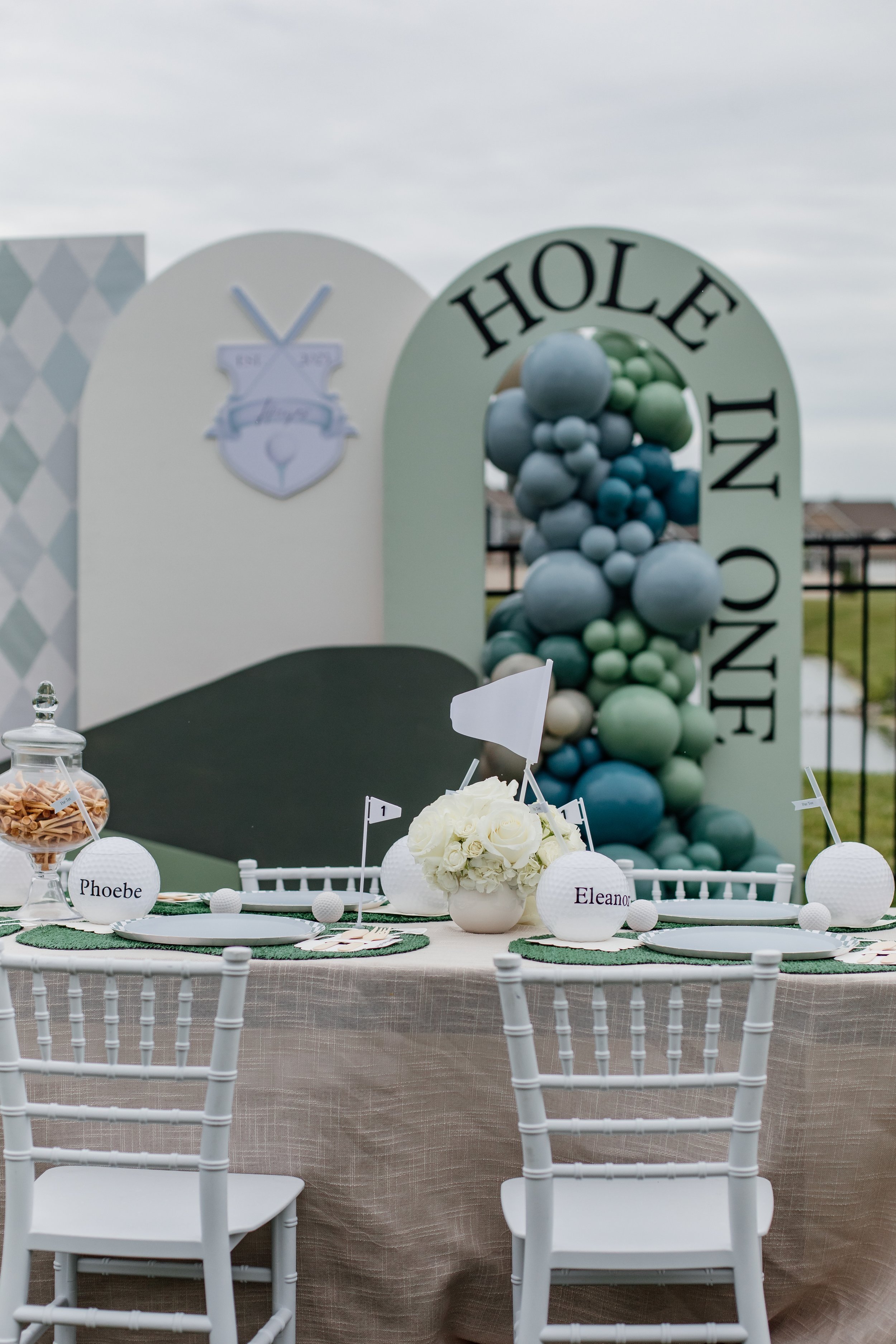 Wedding reception table setup with white chairs, table settings, a centerpiece of white roses, and personalized name cards for guests "Phoebe" and "Eleanor." Background features decorative archways and a balloon display with various shades of blue and green, and a sign with the words "HOLE IN ONE."