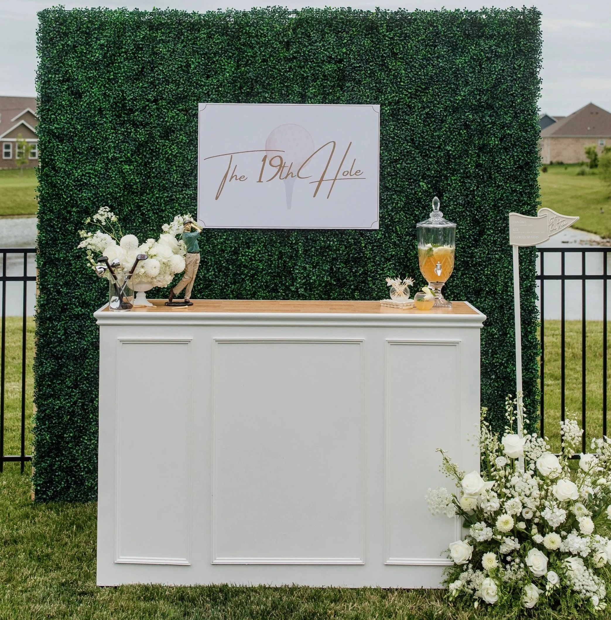 A green hedge backdrop with a white sign reading 'The 19th Hole' in elegant gold script. White floral arrangements and a punch bowl are placed on a white table in front, with some flowers on the grass to the right. The white portable bar is available