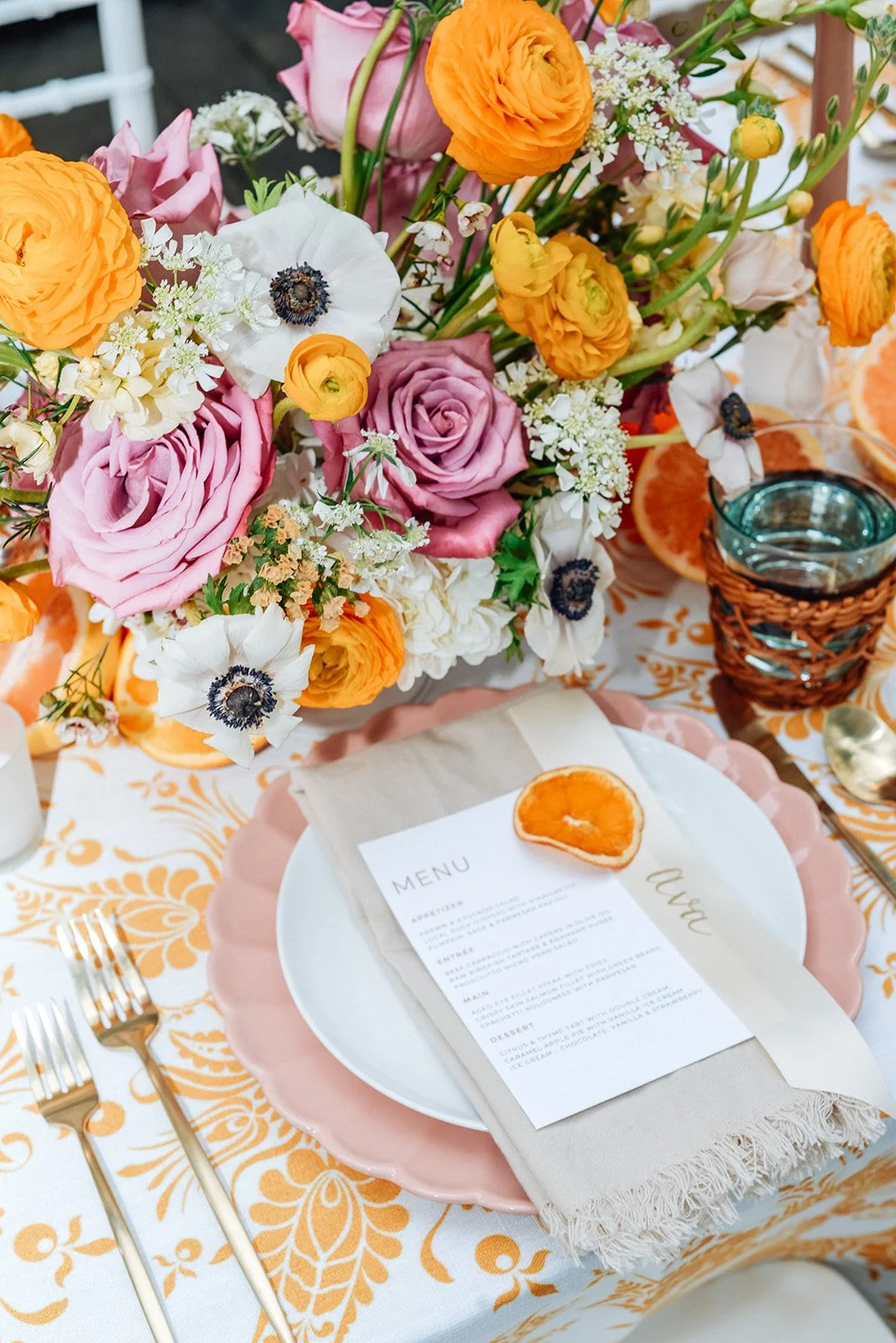 A floral centerpiece with pink, orange, and white flowers on a decorated table set for a meal, including a menu, orange slice, and glass of water.