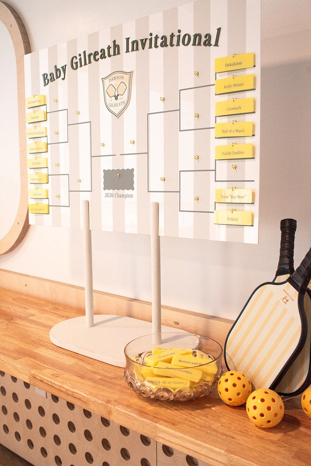 A bracket-style tournament chart titled "Baby Gilreath Invitational" on a wall, with a container of yellow slips of paper, yellow tennis balls, a tennis racket, and a striped bag on a wooden surface.