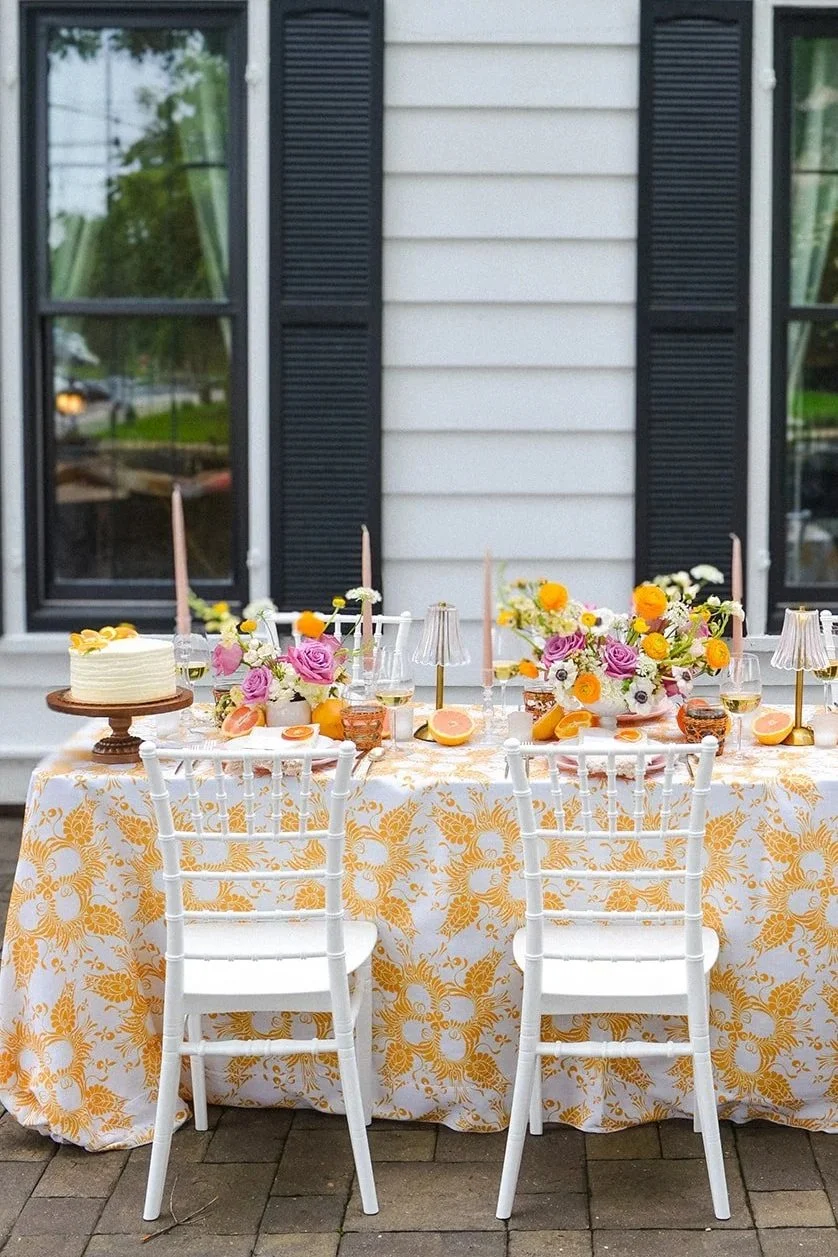 Outdoor table set for a celebration with floral arrangements, candles, a cake, and drinks, in front of a house with white siding and black shutters.