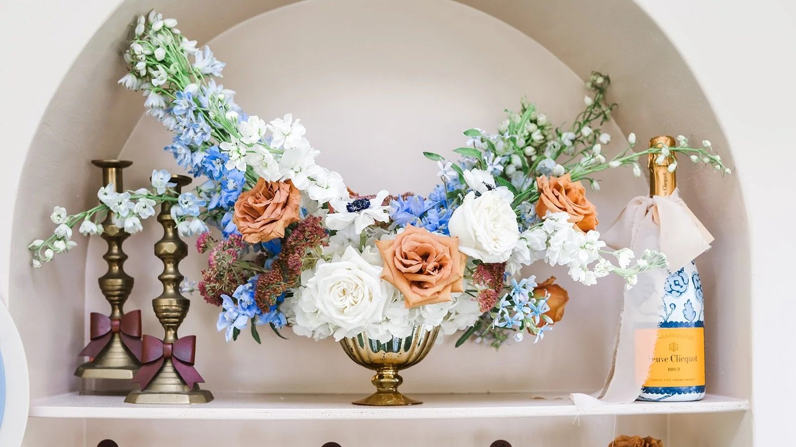 A floral arrangement with white, peach, and blue flowers in a gold vase, flanked by three tall gold candlesticks with purple bows, and a bottle of Veuve Clicquot champagne wrapped with a cloth. These florals were a custom arrangement by Party On Penn