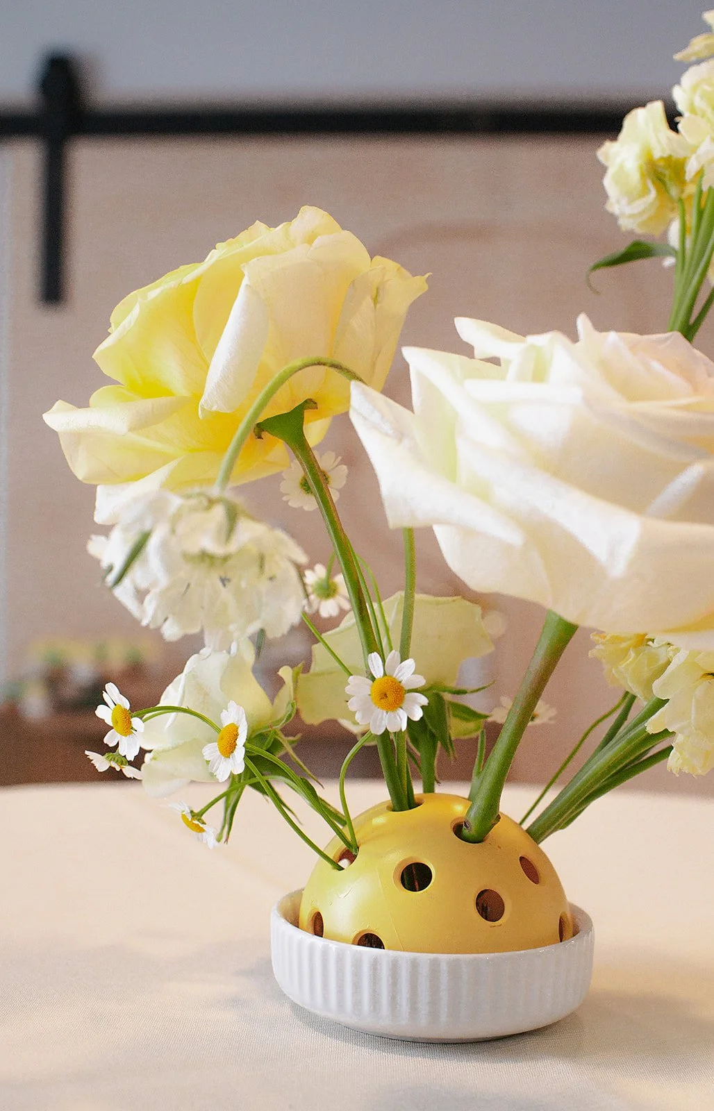 Yellow and white flowers arranged in a decorative yellow vase with large holes, placed in a white bowl on a beige surface.