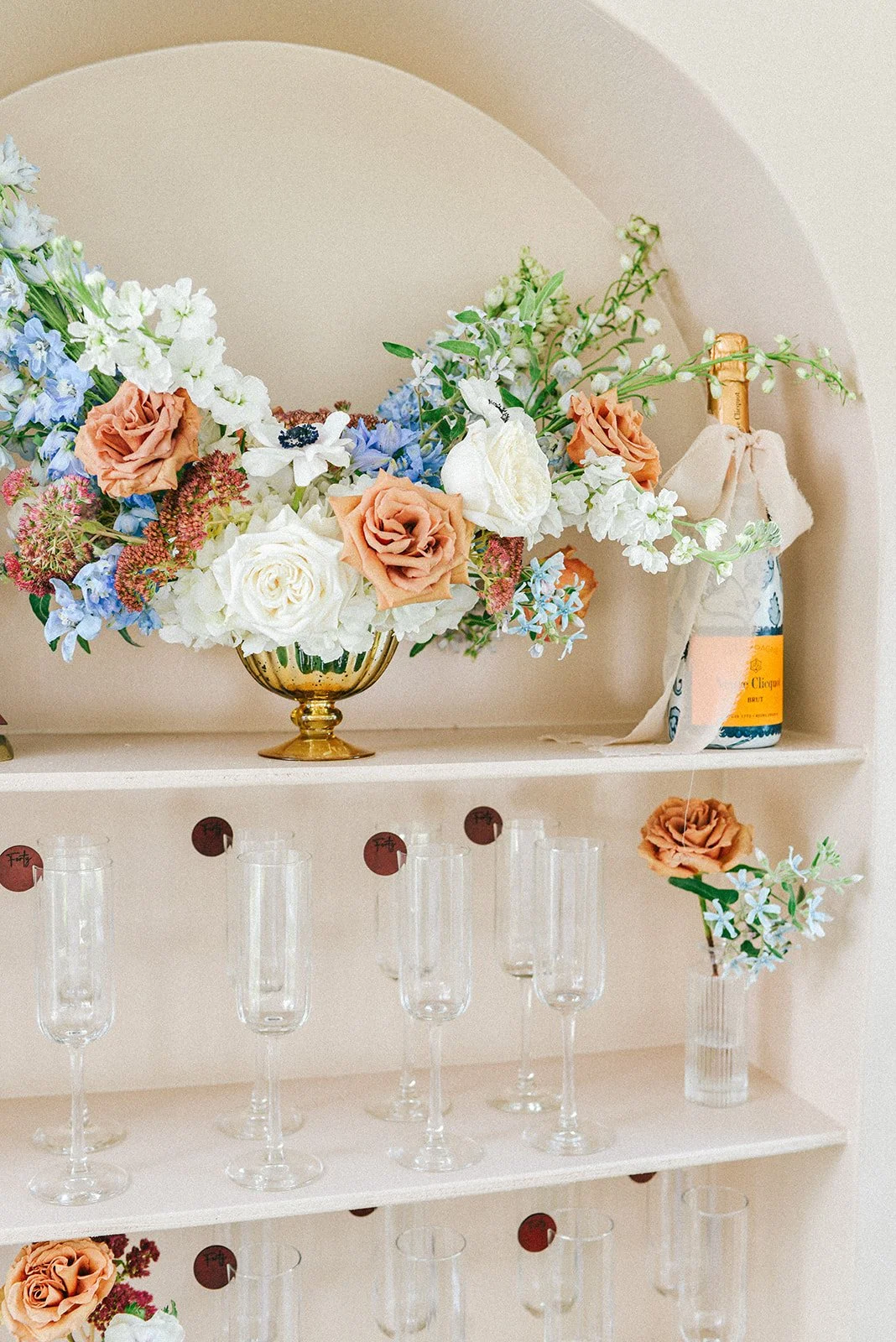 A decorative shelf with a gold vase filled with colorful artificial flowers including roses and other blooms, a bottle with a cloth covering its top, and several empty champagne glasses.