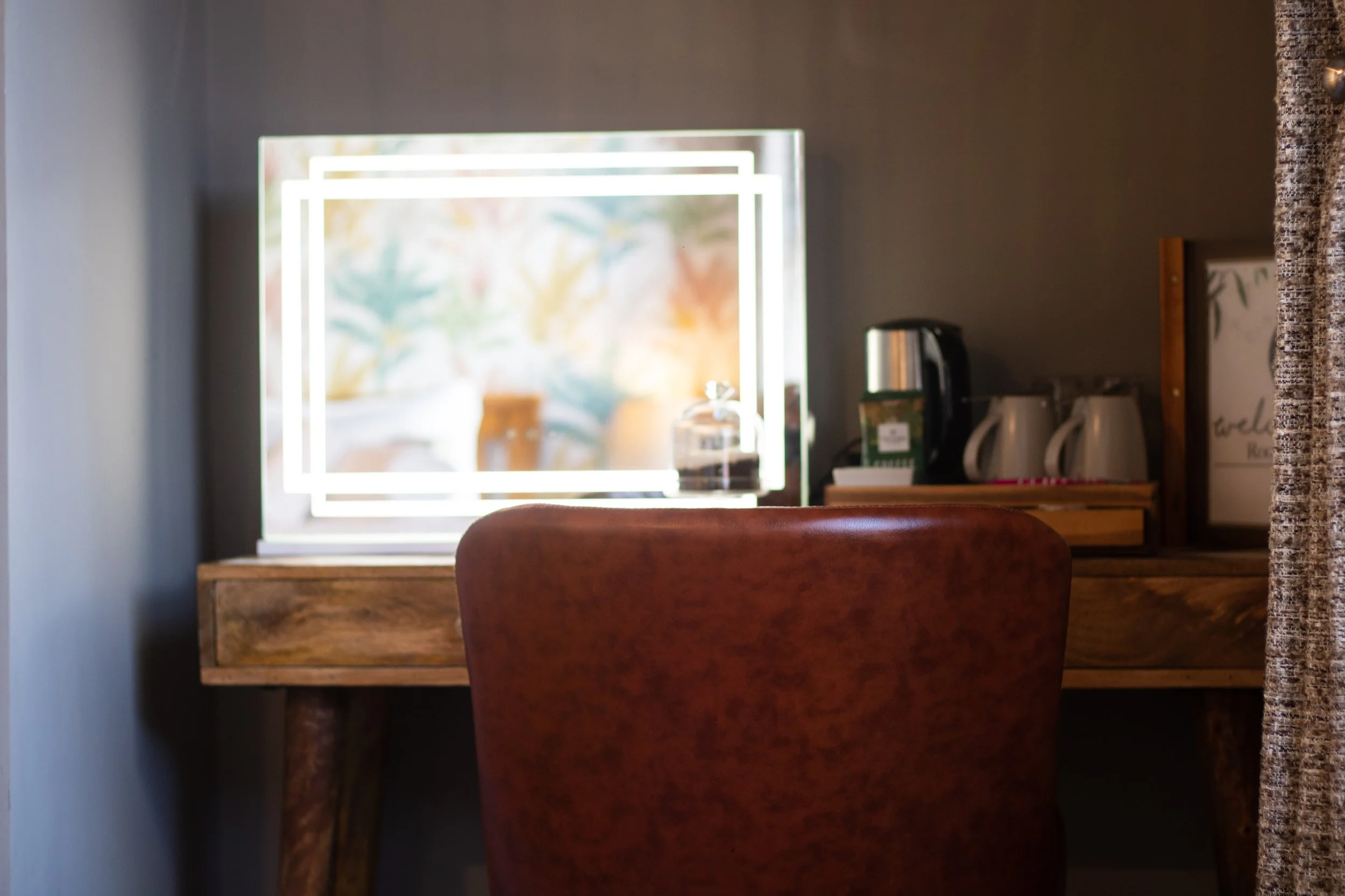 A close-up of a wooden desk with a single brown chair in front of a brightly lit mirror. The background includes a black electric kettle, white mugs, a framed sign, and a curtain on the right.