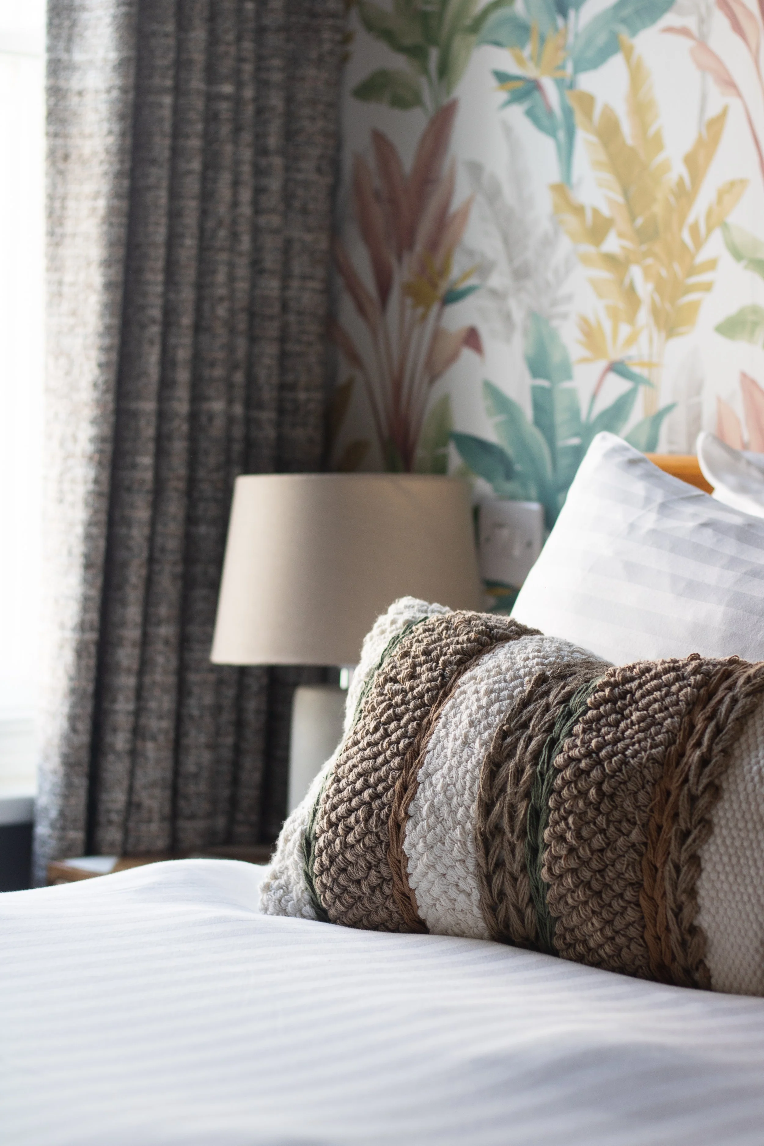 A bedroom with a bed, a decorative pillow, a bedside lamp, and wallpaper with a botanical design.