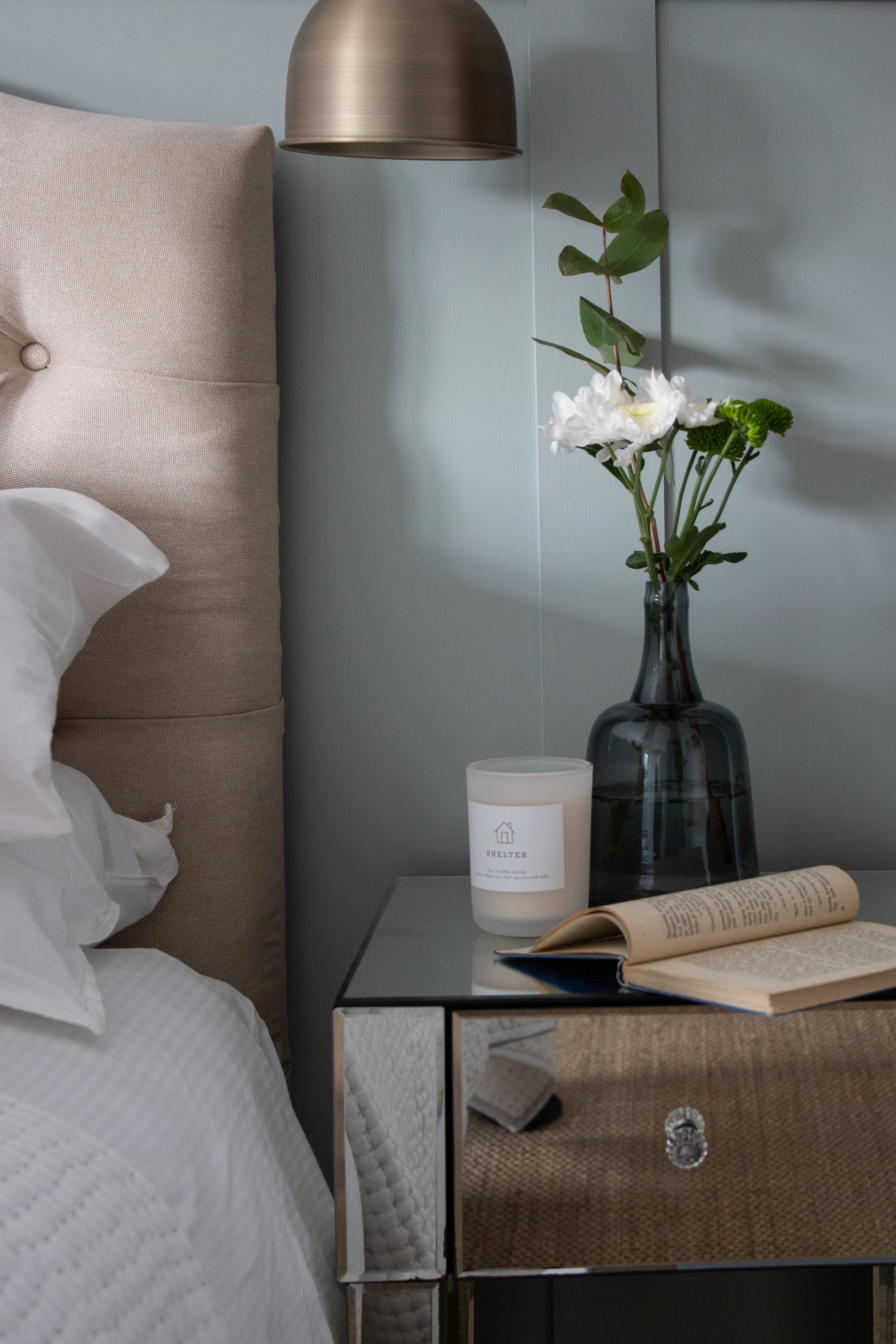 A bedroom nightstand with a black vase containing white flowers and greenery, a candle labeled 'Shelter,' and an open book. Part of a beige upholstered bed with white sheets is visible beside the nightstand. A metallic wall sconce hangs on a light bl