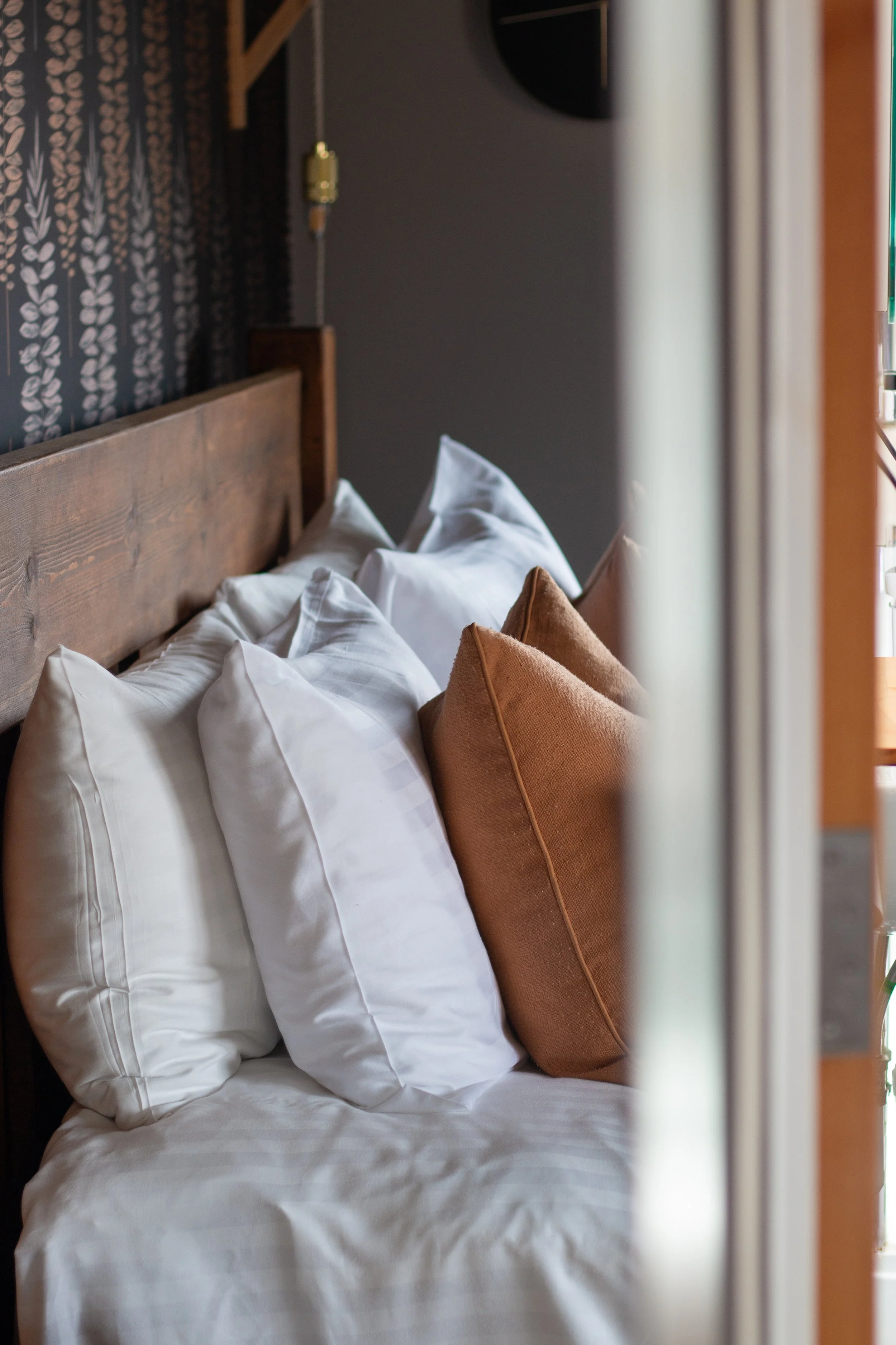 A cozy bed with white pillows and brown accent pillows, against a rustic wooden headboard, in a bedroom with dark walls and patterned wallpaper.