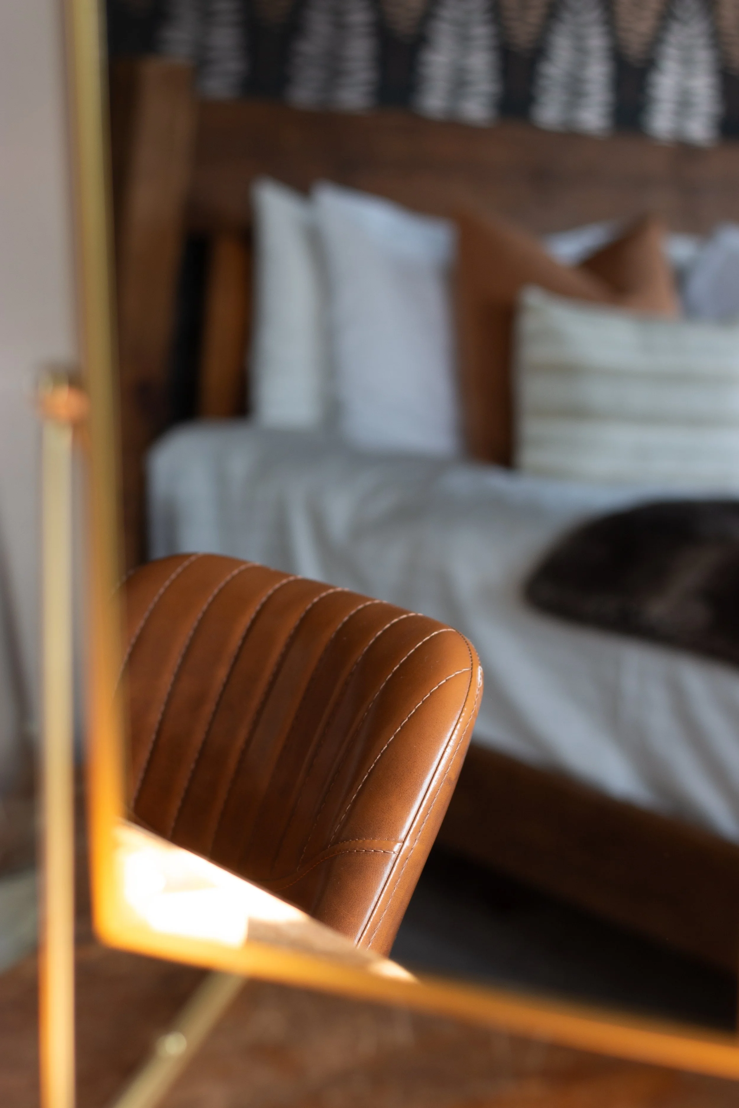 Part of a brown leather chair in foreground with a bed in the background, pillows, and a wooden headboard in a cozy bedroom setting.