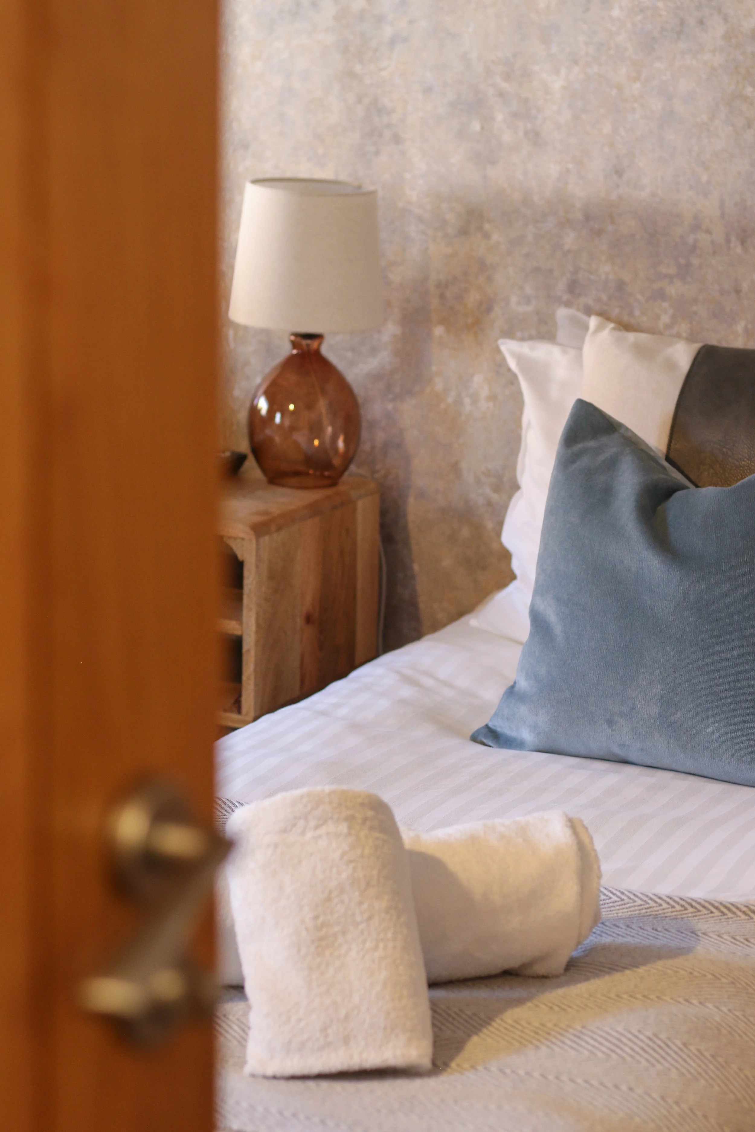 A neatly made bed with white sheets and multiple pillows, including a blue one, with set of rolled white towels on a beige bedspread in a cozy bedroom with textured walls and a wooden nightstand with a table lamp.