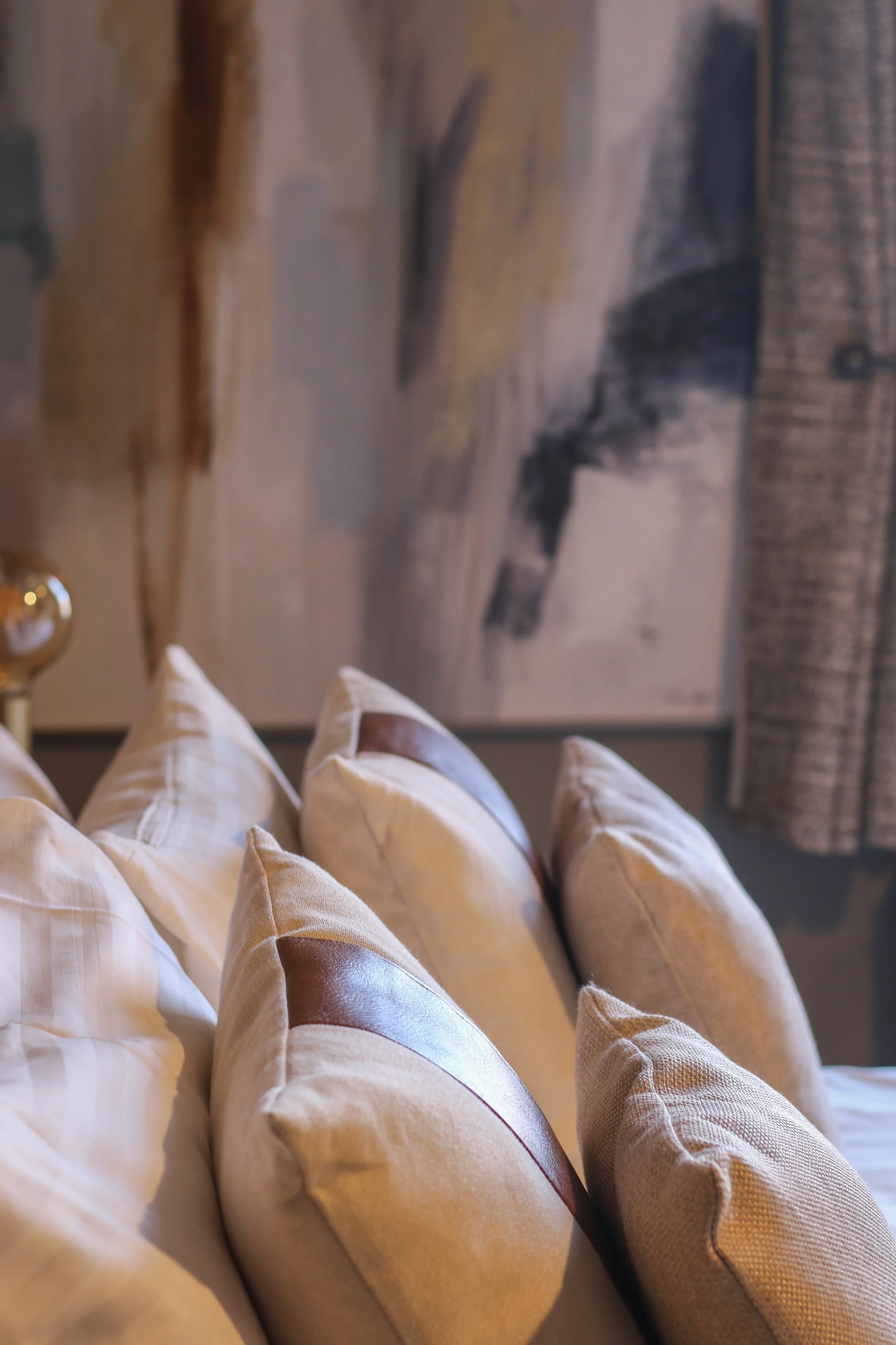 A close-up of beige and brown decorative pillows on a bed with a blurred abstract painting in the background.
