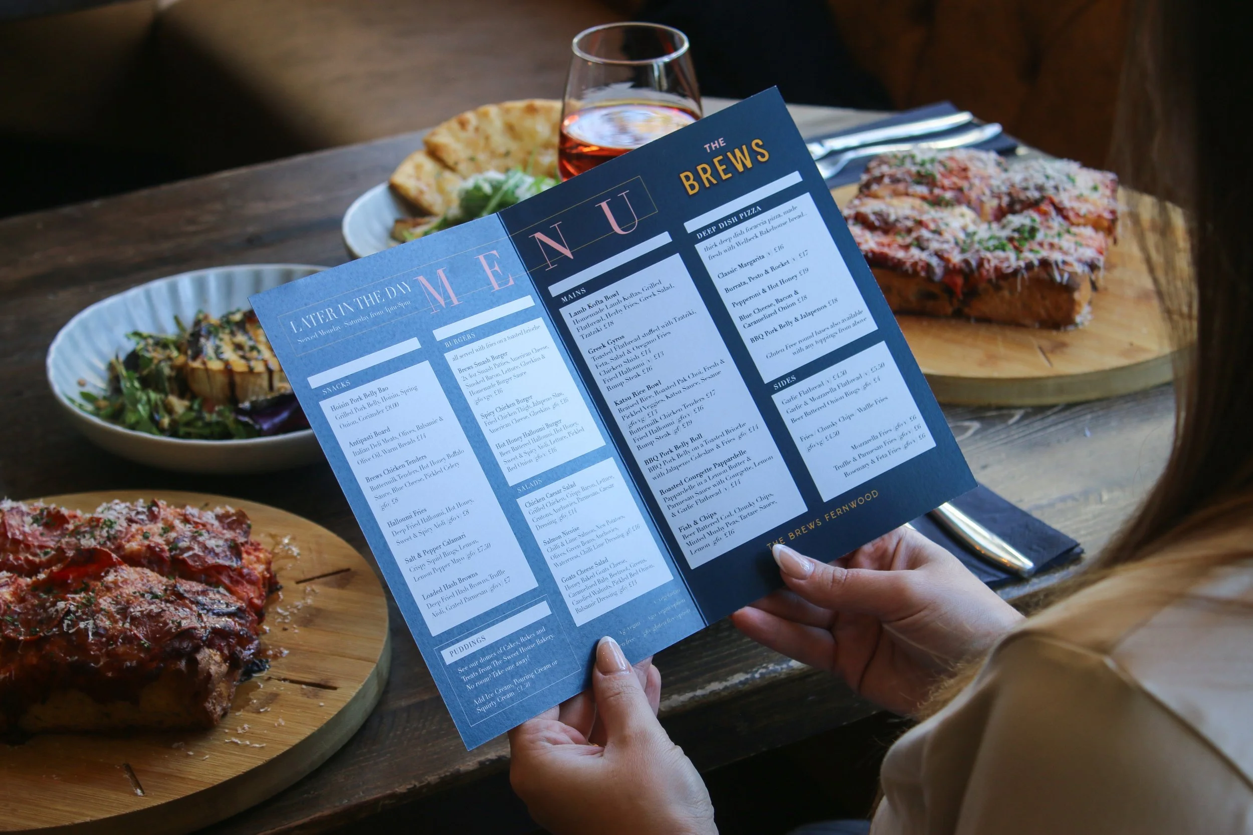 A person holding a blue restaurant menu with food and drink items, sitting at a wooden table with plates of pizza, salad, and a glass of pink beverage.