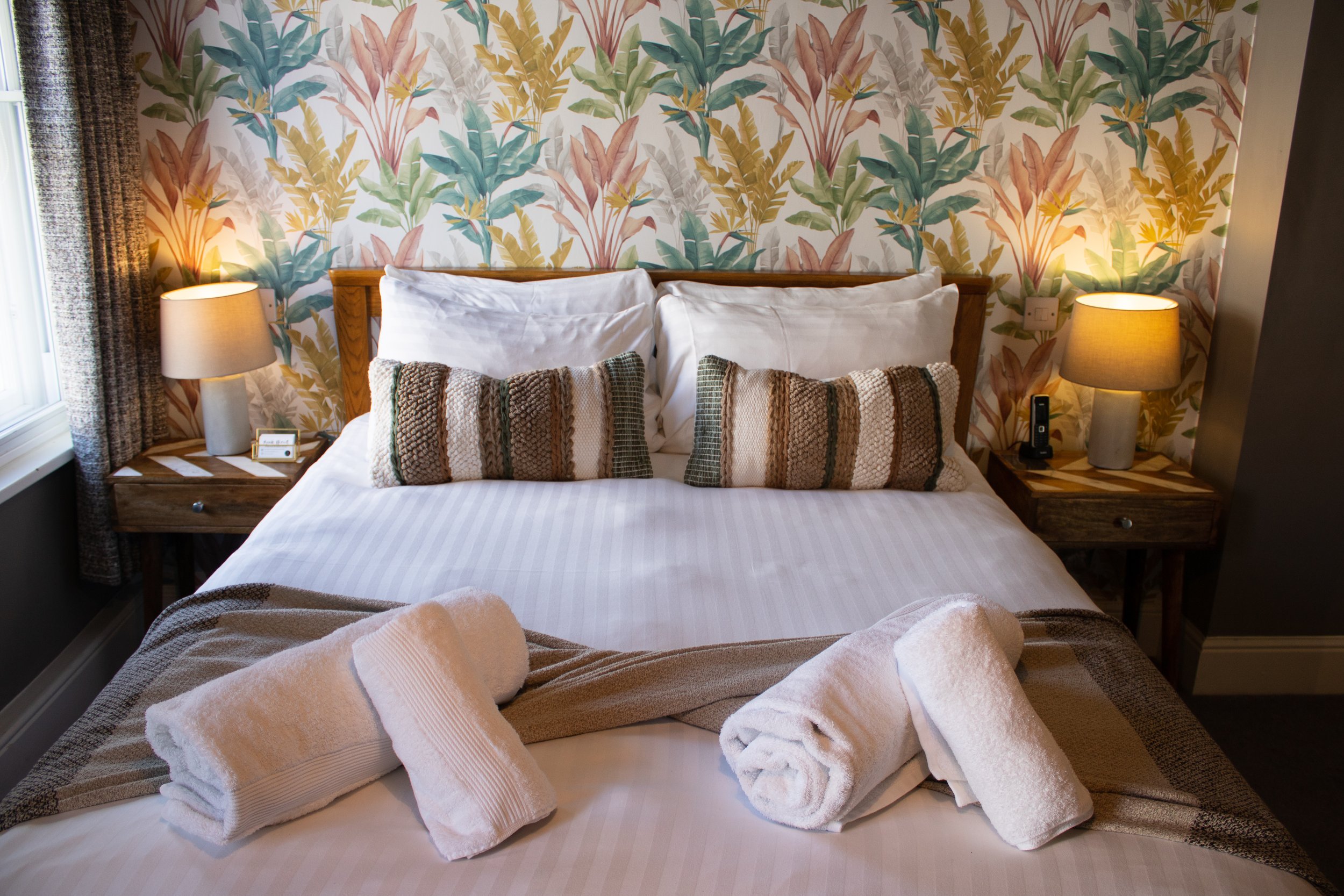 A neatly made bed with white sheets, two white pillows, two decorative striped pillows, and three folded towels, placed in a cozy bedroom with floral wallpaper, two bedside lamps, and wooden nightstands.