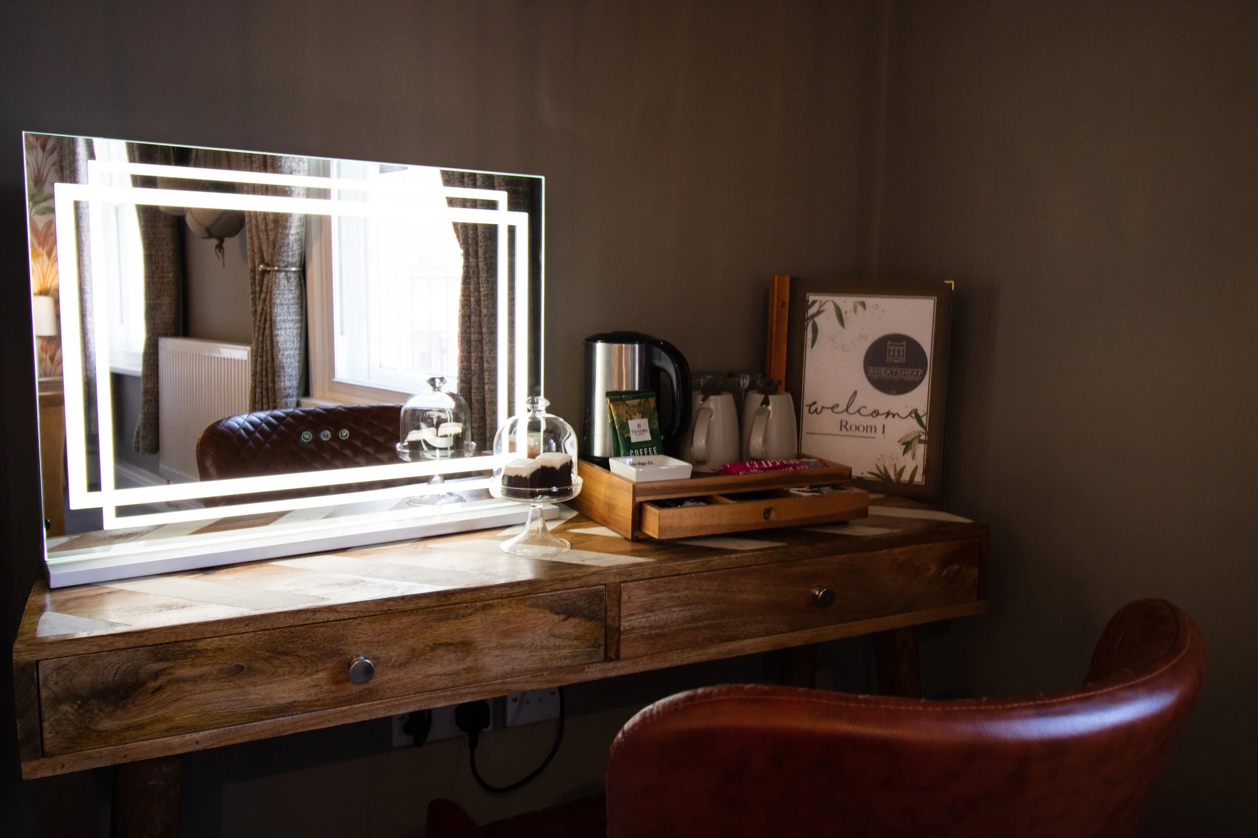 A wooden vanity table with a large illuminated mirror, a glass cake stand with dessert, and a tray with coffee supplies in a cozy room with window curtains.