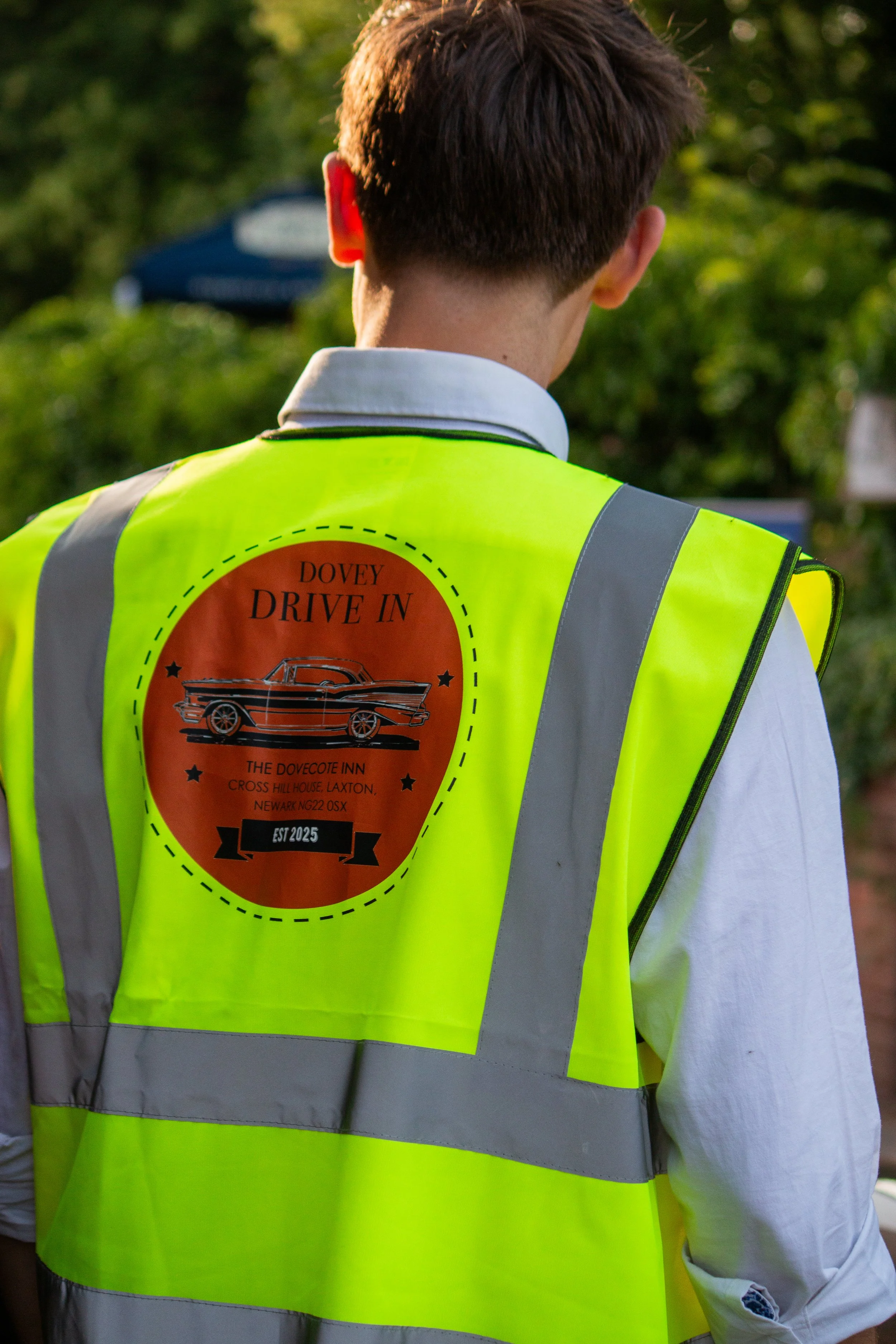 Back of a person wearing a bright yellow safety vest with a logo for the "Dovey Drive In" event, featuring a vintage car and event details.
