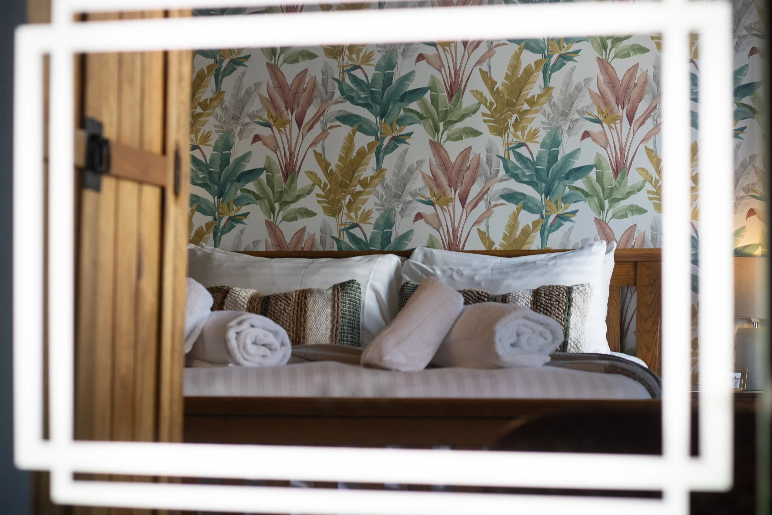 A bedroom seen through a rectangular mirror frame, with a bed decorated with rolled towels and multicolored pillows, and a botanical wallpaper with tropical leaves in the background.