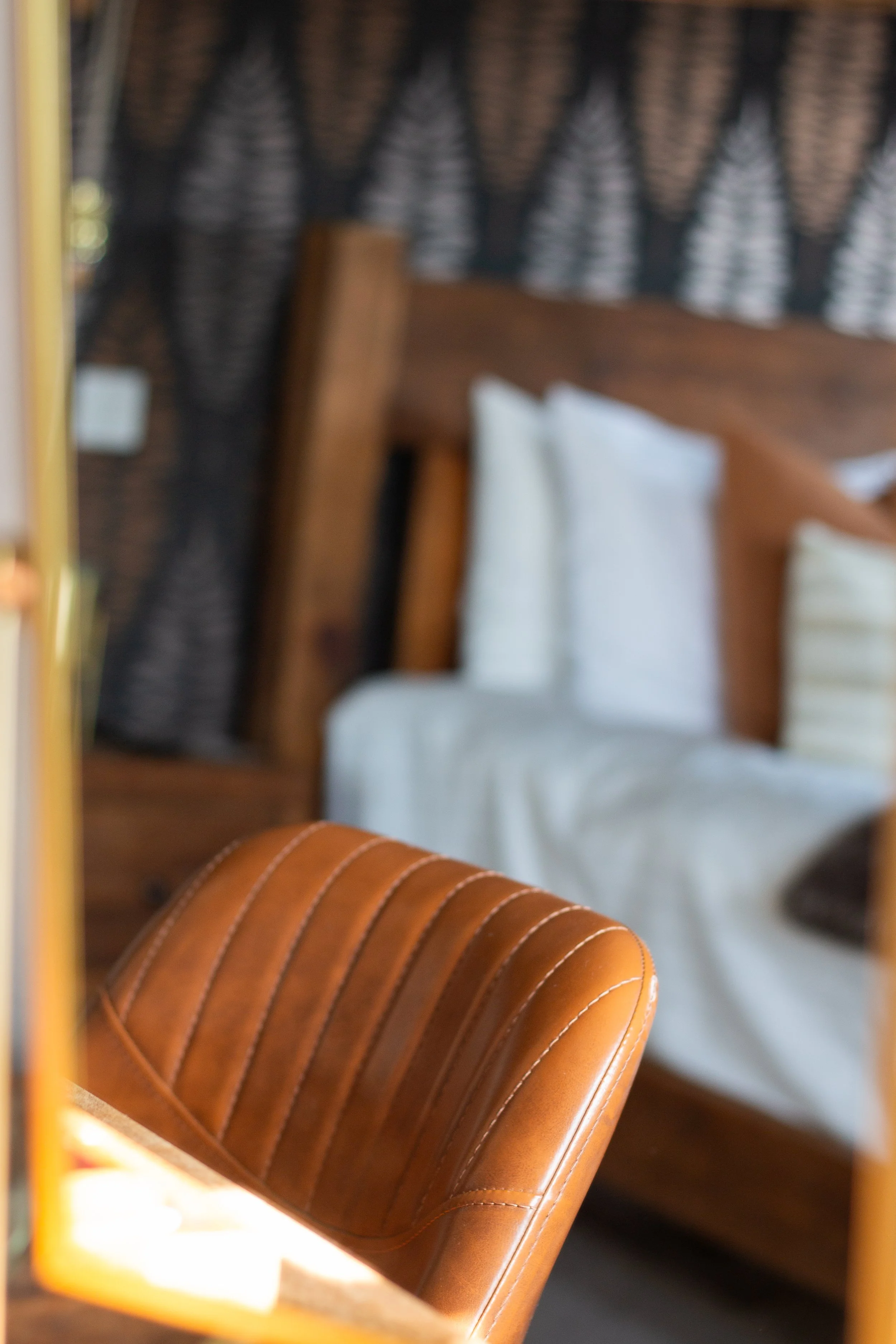 A close-up view of a brown leather chair with a bed and dark patterned curtains in the background.