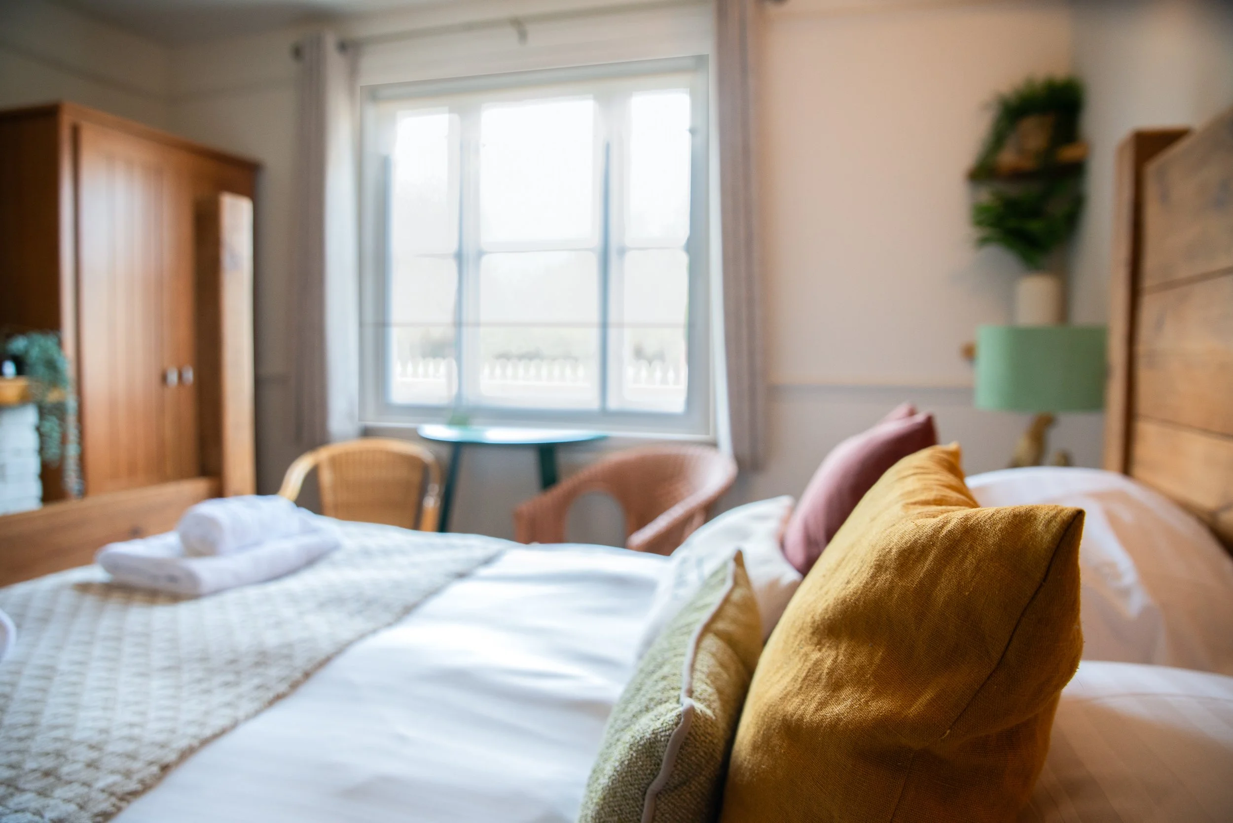 A cozy bedroom with a large bed decorated with yellow and pink pillows, a wooden headboard, and a window with curtains allowing natural light, accompanied by a chair, a small bedside table, and a green lamp.