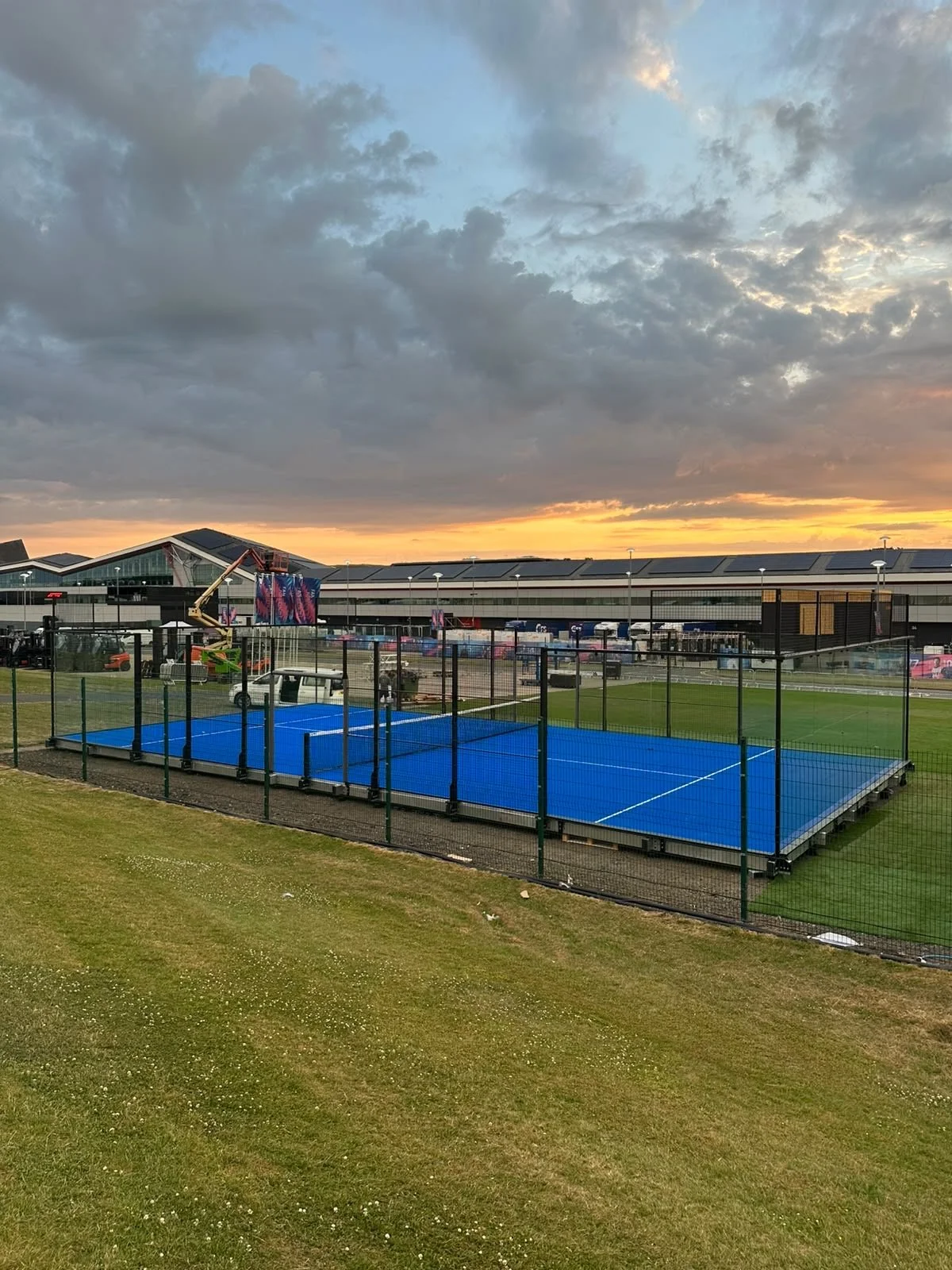 A blue Padel court surrounded by a black wire fence. In the background, there is a building with a modern roof design, a sunset sky with clouds