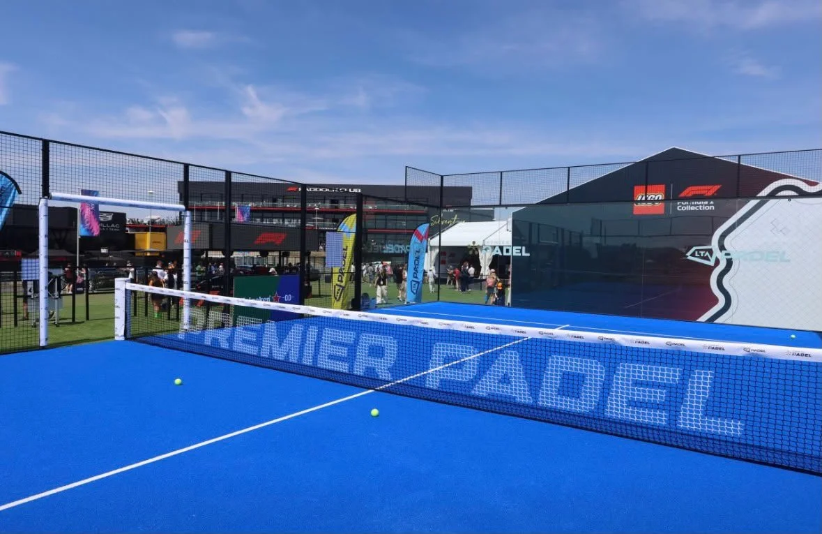 View of a blue padel tennis court at an outdoor sports event, with a black safety net surrounding it, and a few tennis balls on the court at silverstone circuit
