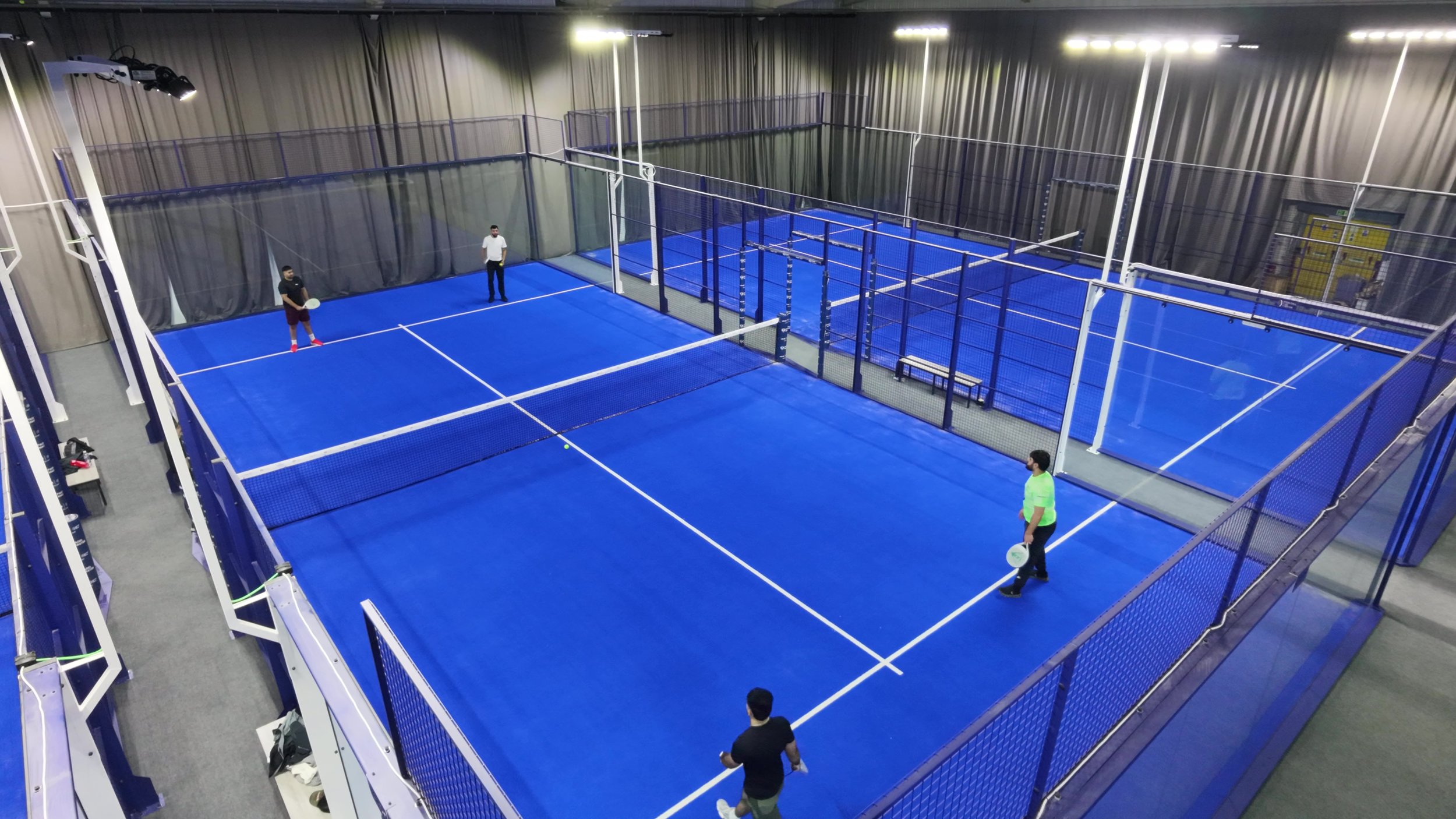 Eight Indoor Padel courts with blue surfaces, netted barriers, and three people playing or observing.
