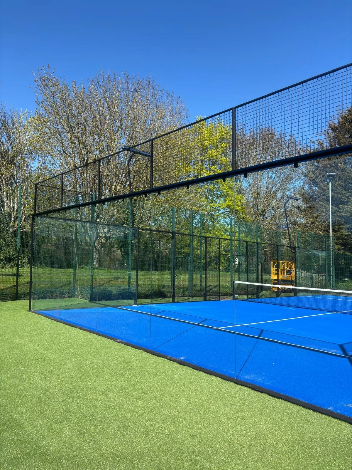 Outdoor Padel court with bright blue surface, surrounded by a black metal fence, green grass, and trees under a clear blue sky.