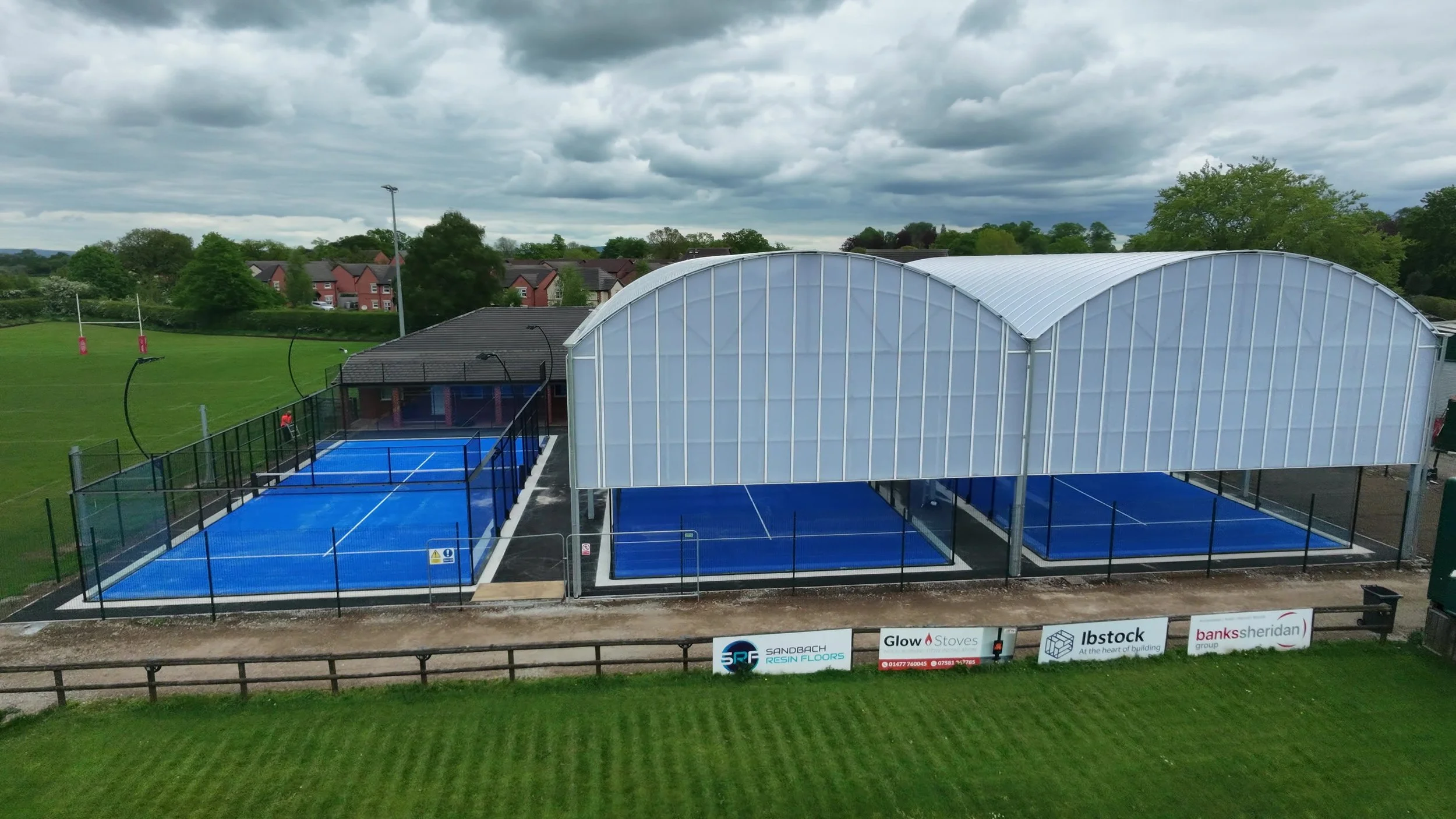 A Padel Complex with 3 Padel Courts with blue turf. Two Padel Courts are underneath a Large white canopy with grass surrounding the site. The Rugby Club in the background