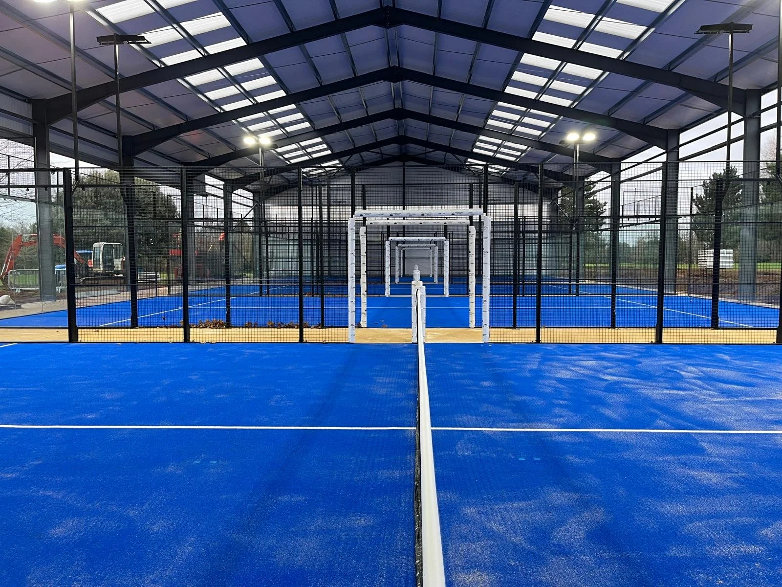 Four Padel Tennis Courts in the UK underneath a large steel canopy. Panoramic MejorSet courts, blue Mondo turf. The canopy is overhead with natural light illuminating the courts. at Brampton Heath Golf Club