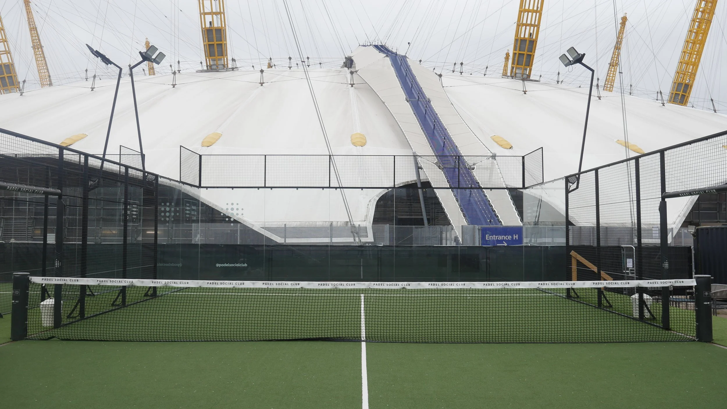 An empty padel tennis court with a green surface, enclosed by a black fence. In the background, there is a large white tent structure with yellow support beams and a blue slide or ramp. There are floodlights outside the court and an entrance sign indicating 'Entrance H'.