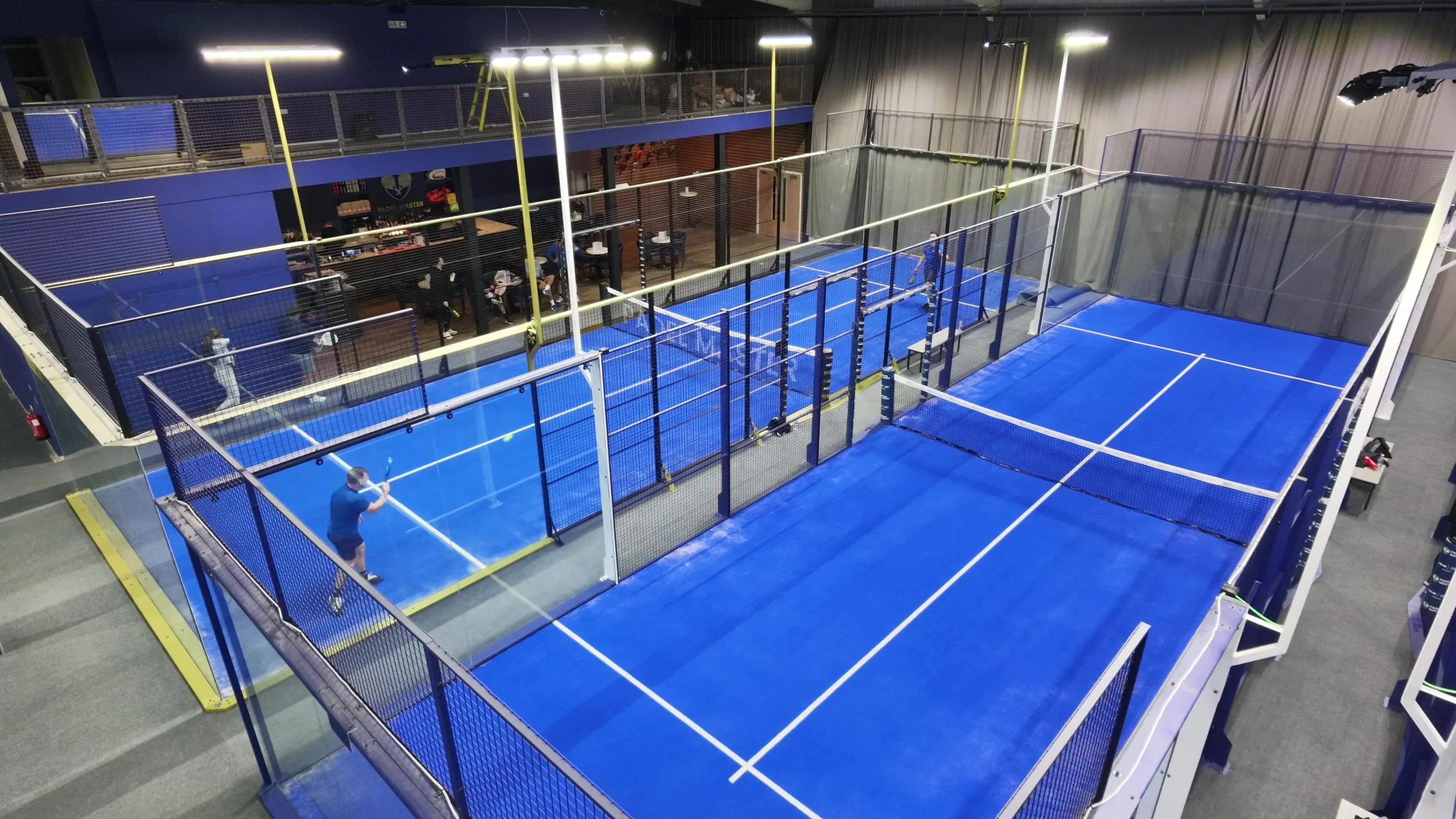 Indoor padel tennis courts with players, spectators, and seating area in the background.