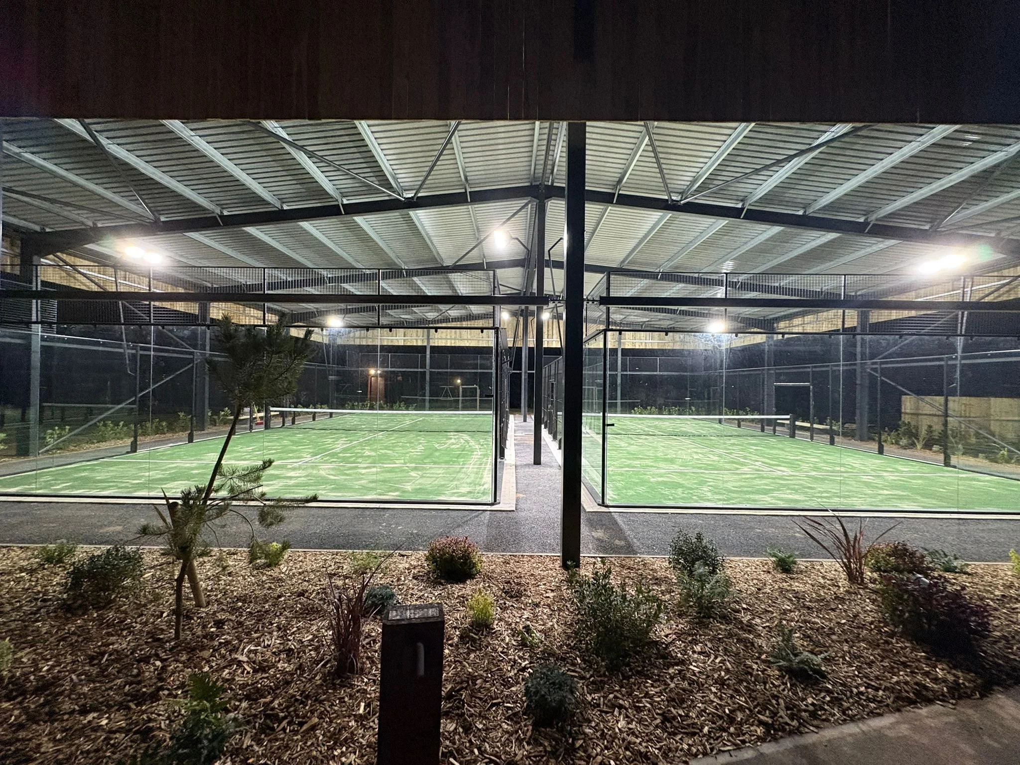 Nighttime view of two outdoor Panoramic Padel Courts under a large Canopy with bright lights, surrounded by a black mesh fence and landscaped with small bushes and a small tree in the foreground.
