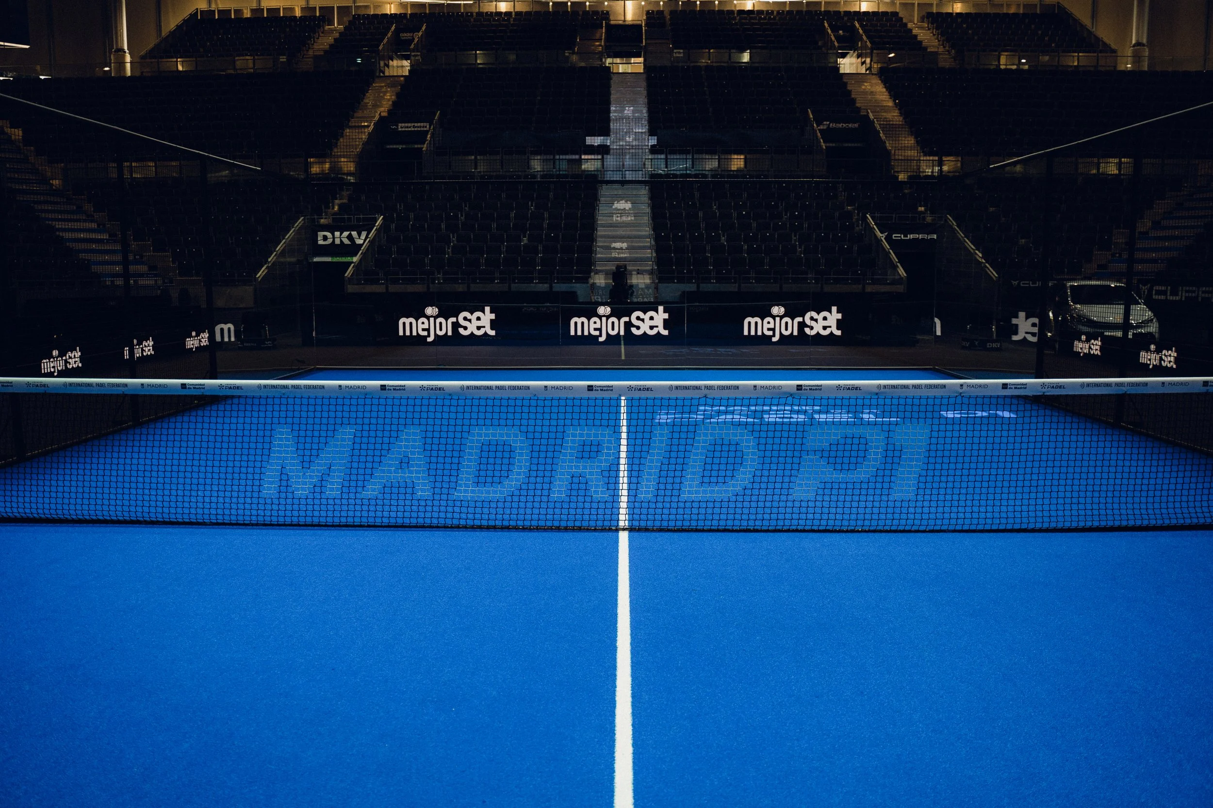 An empty indoor Premier Padel court with blue surface, surrounded by black walls and seating, illuminated by stadium lights.
