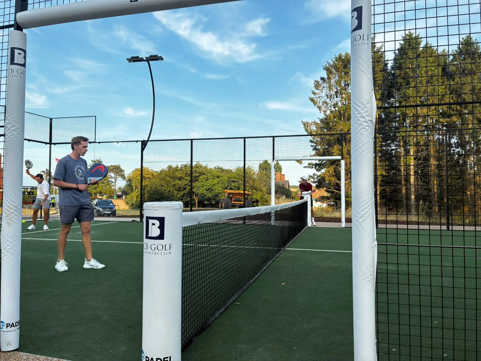 People playing padel on an outdoor court at B Golf Country Club under a partly cloudy sky.