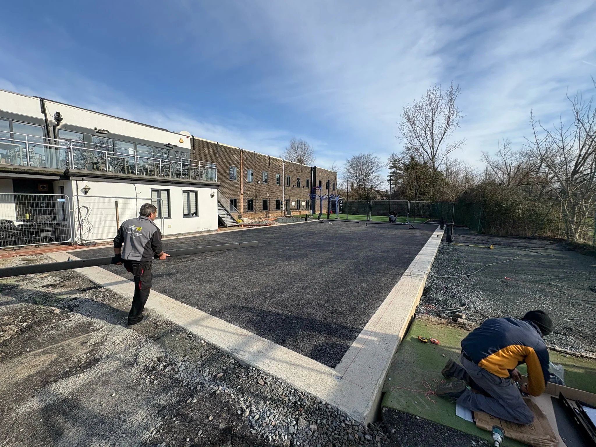 Construction workers installing a new court surface outdoors on a sunny day, with buildings and trees in the background.